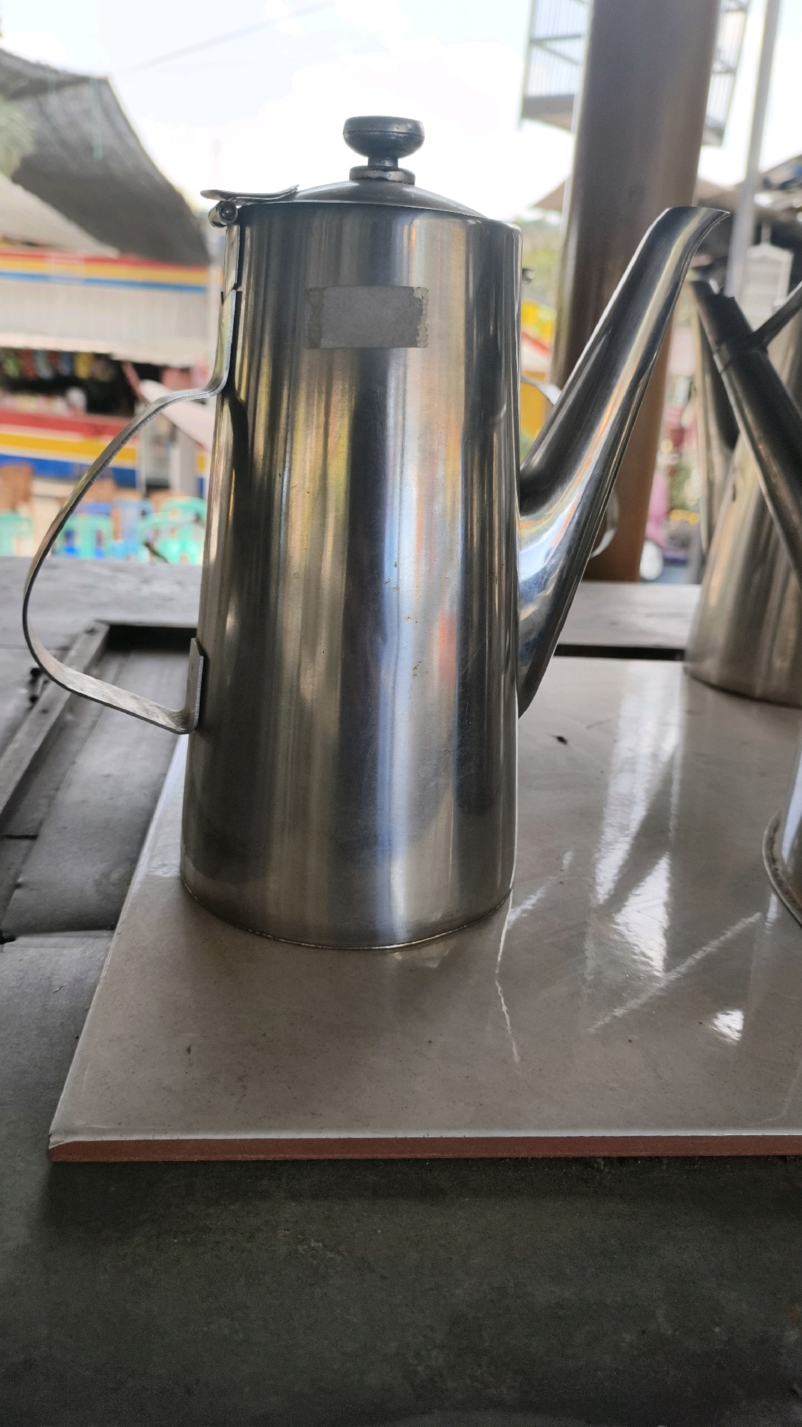 A close-up, eye-level shot captures a metallic teapot with a thin handle and a long spout, resting on a square, light-colored tile. The teapot is silver and reflects the surroundings, including a blurred background of colorful stalls and structures under a bright, hazy sky, suggestive of an outdoor market or roadside establishment in Air Paoh, Indonesia. The tile is positioned on a darker, textured surface, possibly a table or counter. Another metallic object, similar to a teapot, is partially visible to the right of the main subject. The overall scene is well-lit, with the light reflecting strongly off the metallic surfaces.