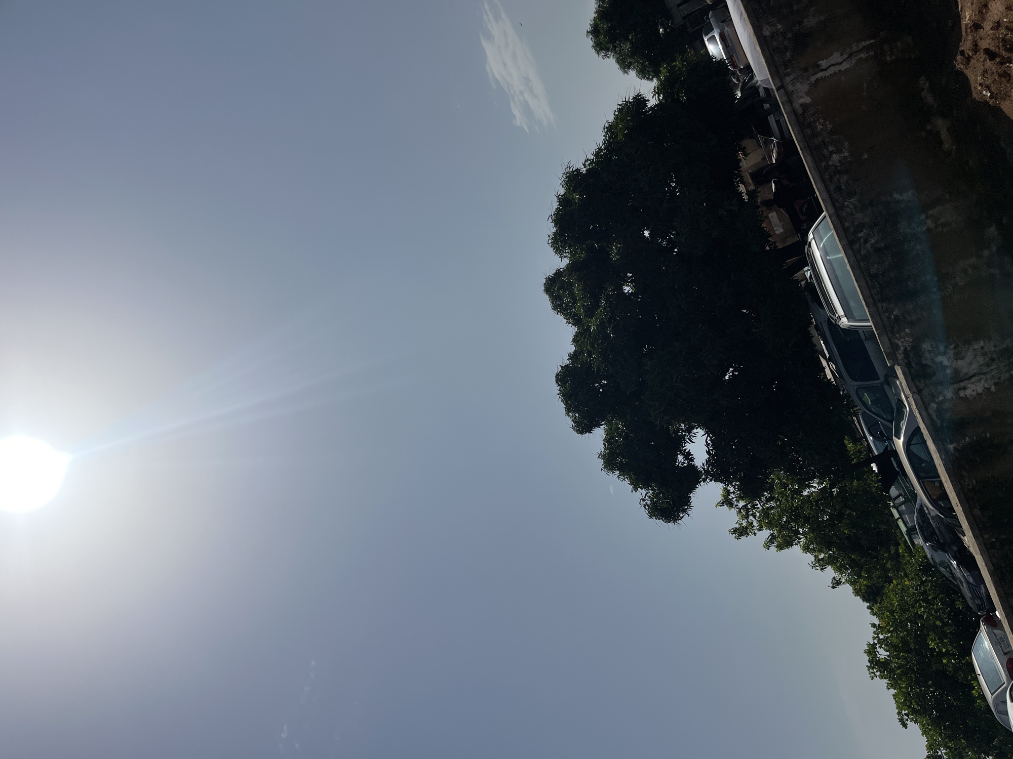The image, captured in Bukuru, Nigeria, depicts a bright, sunny day with the sun prominently displayed in the upper left quadrant of the sky, casting strong lens flare. The sky is a pale blue, with faint clouds visible. In the foreground, a weathered concrete wall runs diagonally across the bottom right of the frame. Behind the wall, a collection of vehicles, including cars and what appears to be a van or SUV, are parked. Lush green trees are abundant throughout the scene, with a large, dense tree with dark green foliage dominating the center of the image. The overall impression is that of an outdoor area, likely a parking lot or an open space adjacent to buildings, under a strong sun.