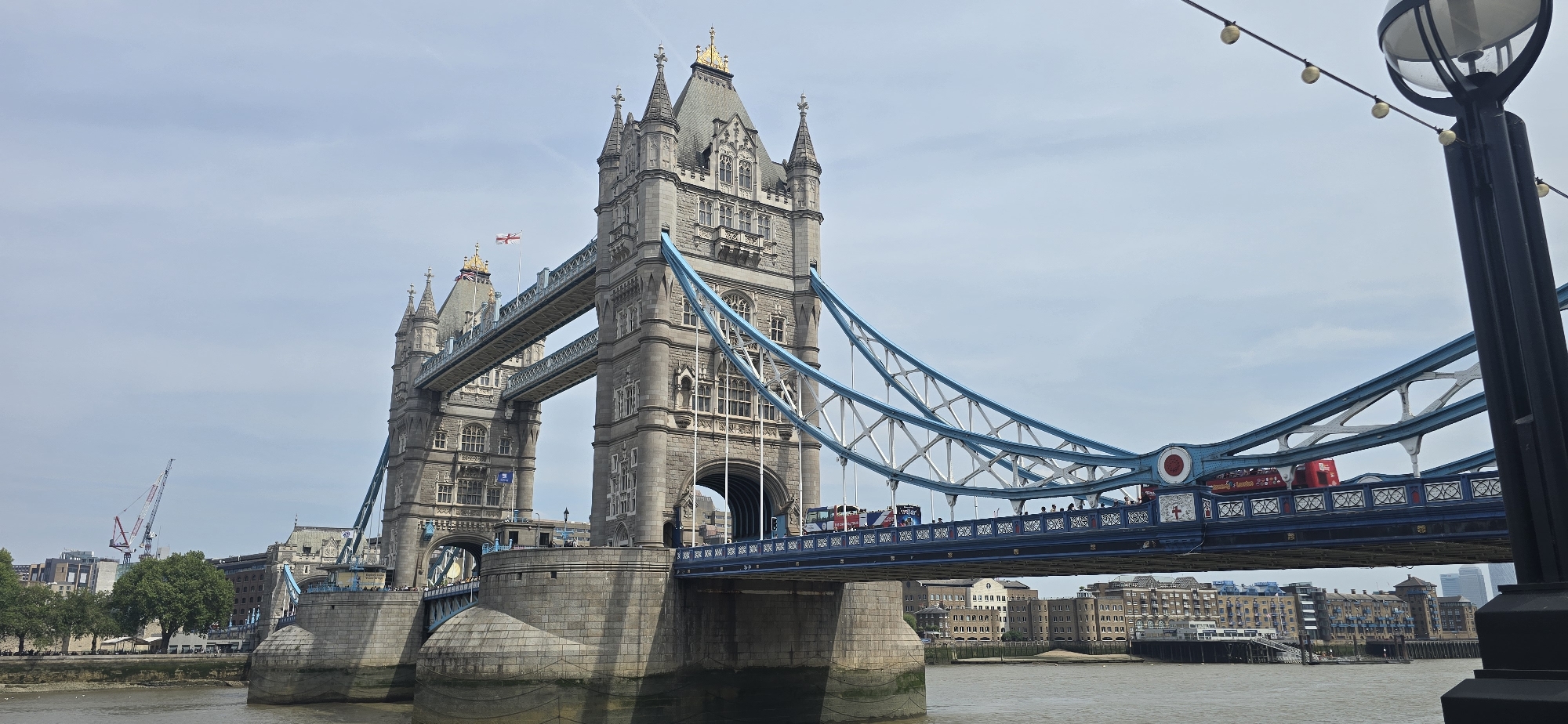 The image shows the Tower Bridge in London, United Kingdom. The bridge is in the foreground, with its two towers visible, along with the blue and white suspension structure. A red double-decker bus and people are on the bridge. The River Thames is in the foreground, and buildings can be seen on both sides of the river in the background. A black lamppost is on the right. The sky is overcast. A construction crane is visible in the distance on the left.