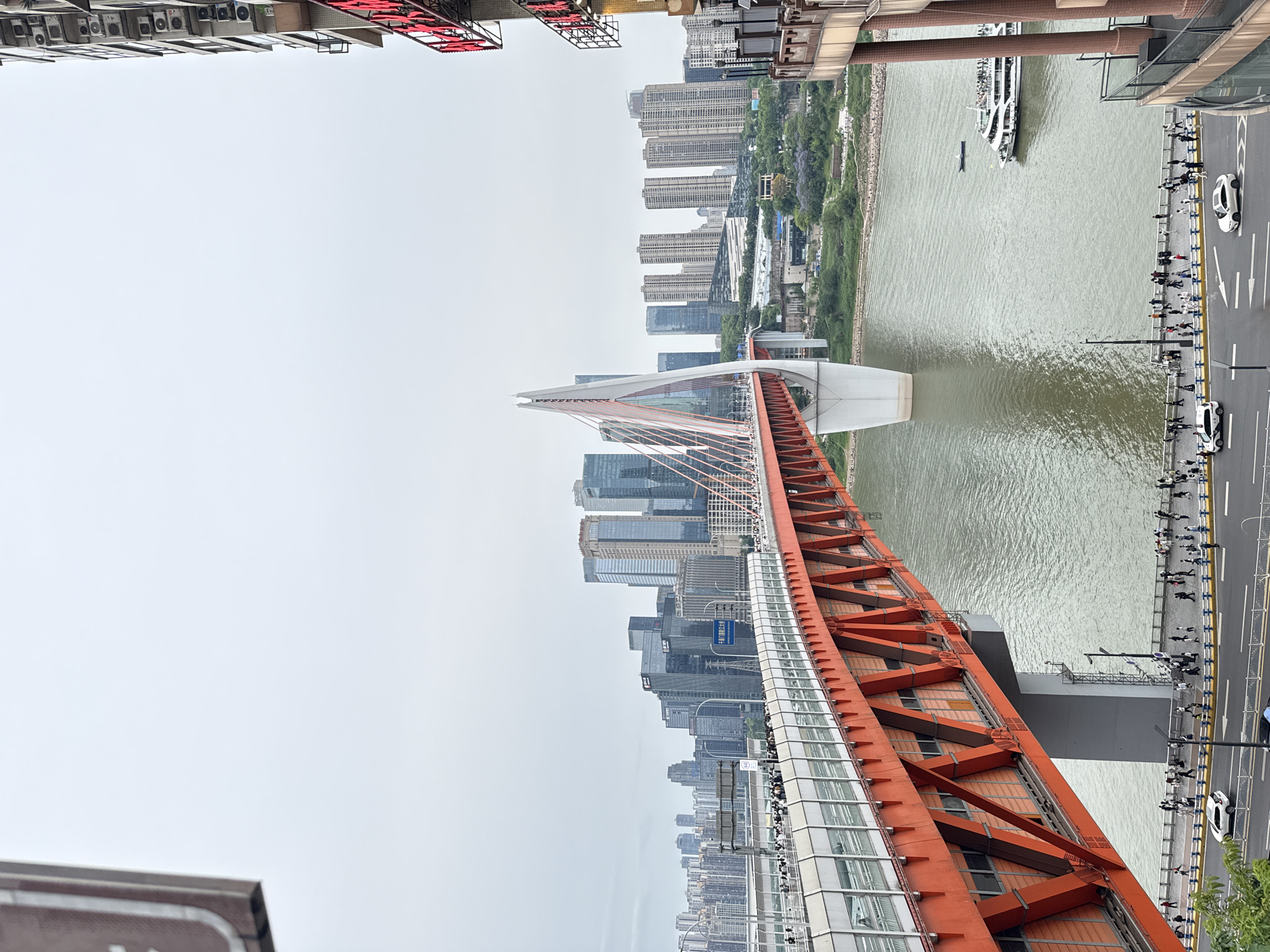 The image shows an elevated view of the orange-colored, multi-span Chaotianmen Bridge, spanning a wide river, likely the Yangtze River in Yuzhong District, Chongqing, China. On the right side of the bridge, a pedestrian walkway lines the river with numerous people walking. Cars are visible on a road parallel to the walkway. A river cruise boat and other vessels are on the river. In the background, numerous high-rise buildings form a dense urban skyline partially obscured by overcast skies. Buildings and objects are visible on the balcony/top of a building where the photograph was taken.