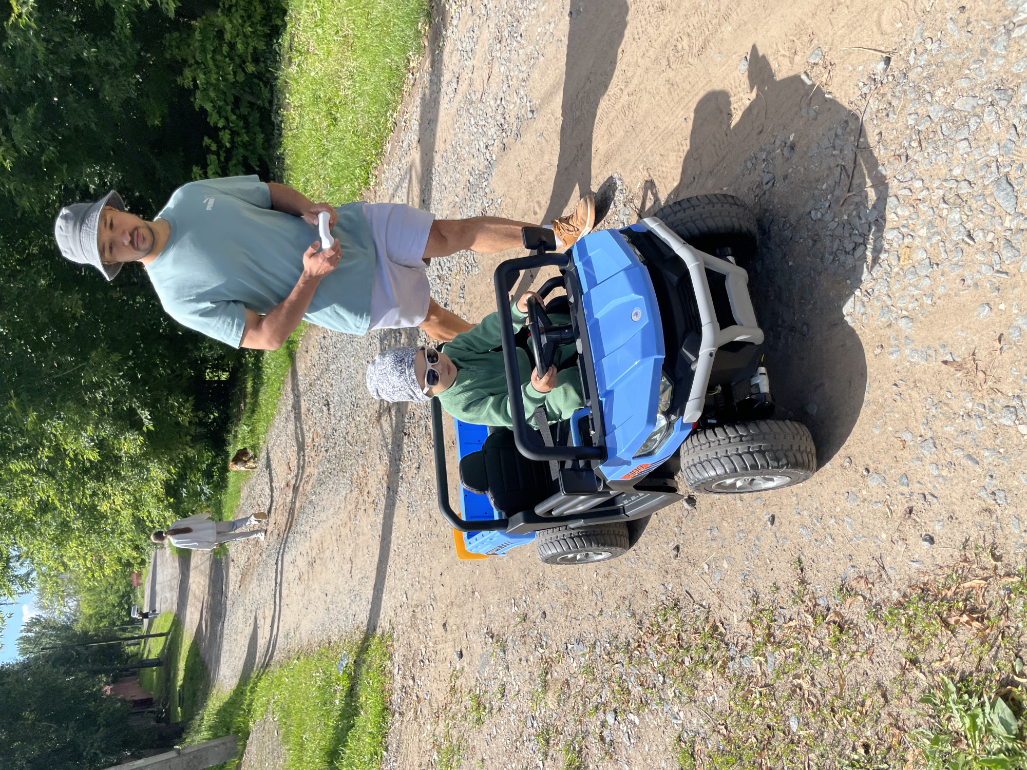 An adult male wearing a light blue t-shirt and light-colored shorts stands on a gravel path, holding a white remote control. Beside him, a child wearing a green hooded jumpsuit and a grey patterned beanie with white sunglasses sits in a blue toy electric car. The car has black trim and oversized tires. In the background, a young girl in a light grey sweatsuit walks away from the camera, and a brown dog is visible on the grass to her right. The path is lined with trees and grass, with a brick wall visible on the left side of the frame. The scene appears to be an outdoor, rural or suburban setting.