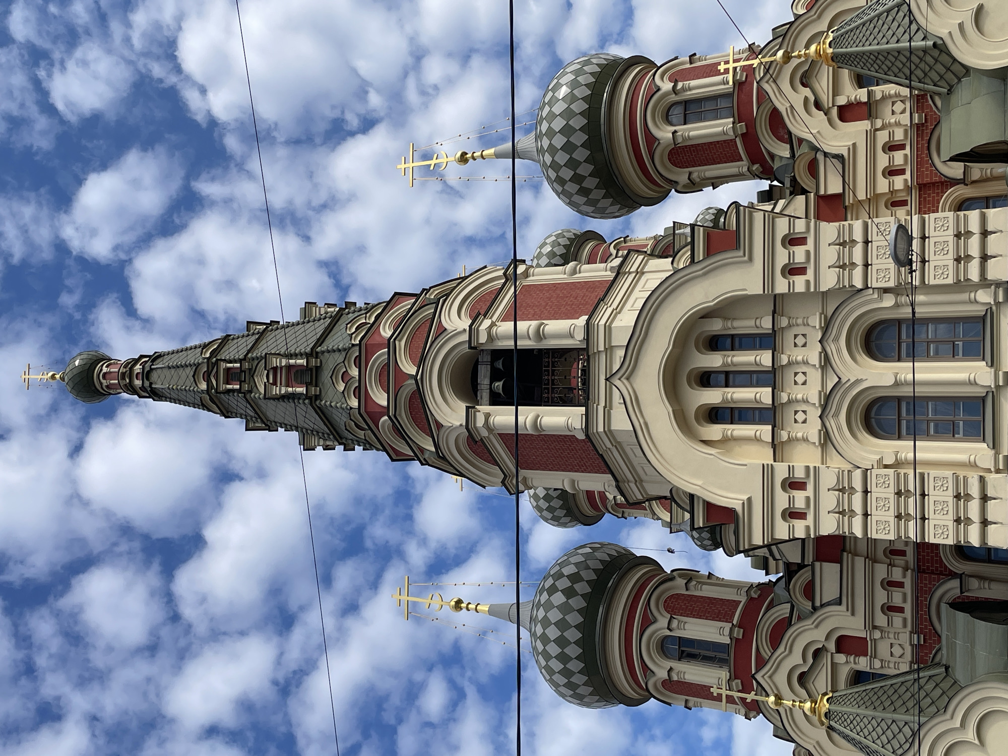 The image displays a majestic and ornate Russian Orthodox church building, captured from a low-angle perspective against a vibrant blue sky dotted with fluffy white clouds. The primary subject is the church's intricate architecture, characterized by a towering, multi-tiered bell tower or spire that reaches high into the sky. This central spire, like the other prominent domes, is topped with a golden Orthodox cross.
The church's facade is a rich blend of traditional design, featuring sections of red brickwork interspersed with elaborate cream or light beige decorative elements. These details include arched window frames, ornate cornices, and detailed relief patterns. Several onion domes are visible, some exhibiting a distinctive checkered pattern in hues of gray-green and light gray, each also surmounted by a golden cross. Slender chains or wires are visibly attached to some of the golden crosses, anchoring them to the domes.
The bright, natural lighting indicates it is daytime, and the clear sky with scattered clouds suggests fair weather. There is no specific activity or event taking place; the image is a static architectural study. Thin, dark cables or power lines traverse the frame, crossing over the church and the sky. No visible text is present in the image. Given the architectural style and provided context, the church is located in Saint Petersburg, Russia.