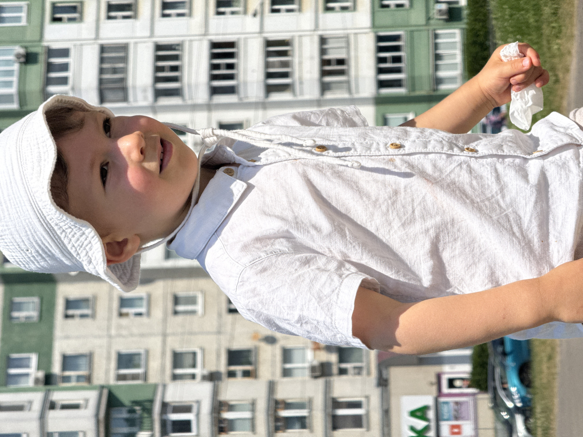 A young boy wearing a white bucket hat and a white collared shirt stands in front of a tall, multi-story apartment building. The building has a green and grey facade with numerous windows. The boy is looking to his right, with his mouth slightly open. His right arm is bent, and he is holding a crumpled piece of white tissue in his hand. In the background, out of focus, there is a street with cars and a store with a green "KA" sign. The lighting suggests it is daytime, with sunlight casting shadows on the boy.