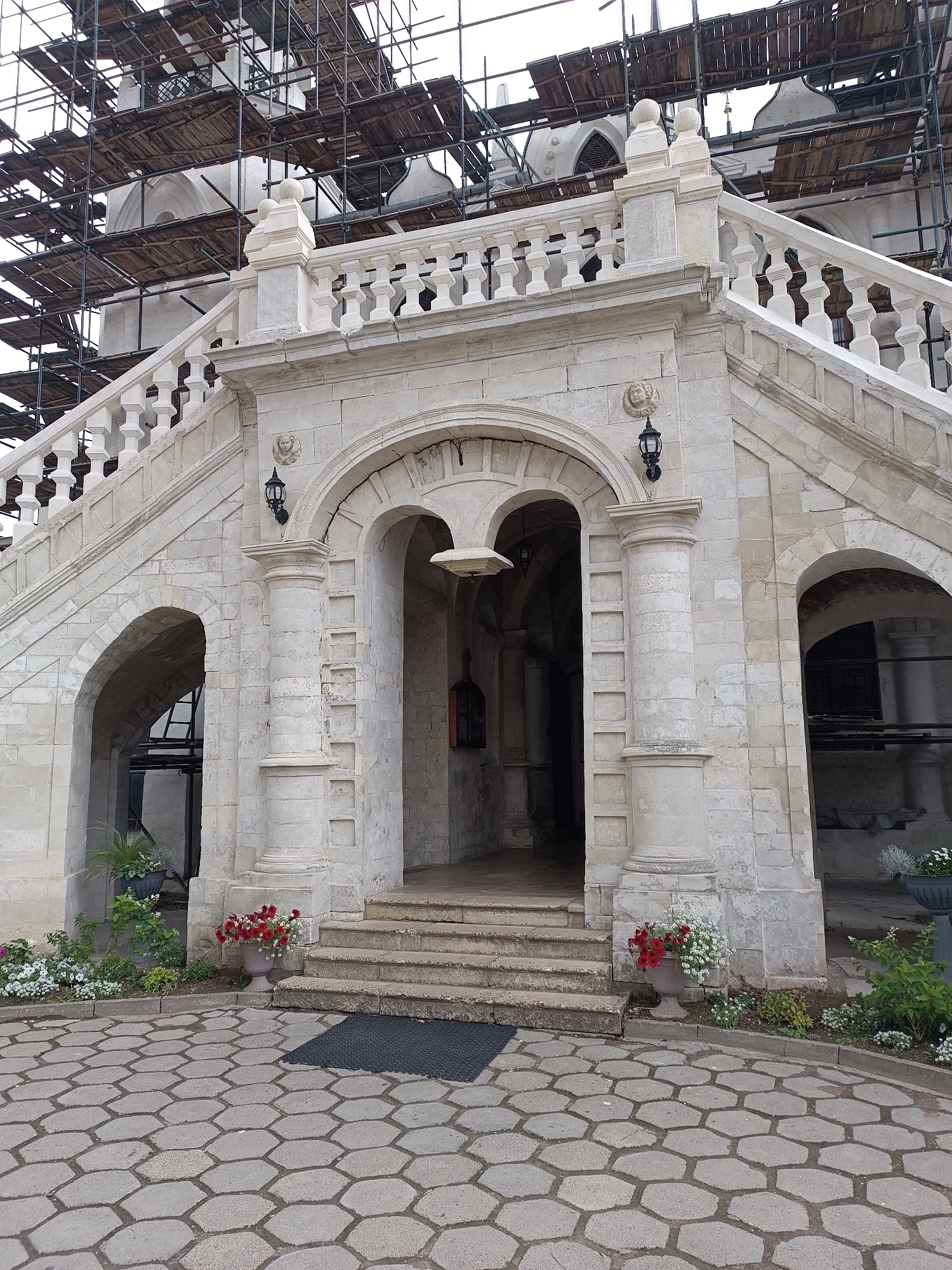 The image depicts the stone staircase and entrance of the Church of Vladimir Icon of the Mother of God in the village of Bykovo, Russia. The church is undergoing renovation, evidenced by the extensive scaffolding covering its upper sections. The foreground features a hexagonal paved courtyard, with a dark mat placed in front of the steps. To the left and right of the entrance, there are large stone planters filled with red and white flowers. The staircase itself is constructed of weathered stone, with a white balustrade lining its upper sections. The entrance is framed by a large archway supported by columns, with two smaller arched openings on either side. Decorative elements, including a carved face above a lamppost, are visible on the facade.