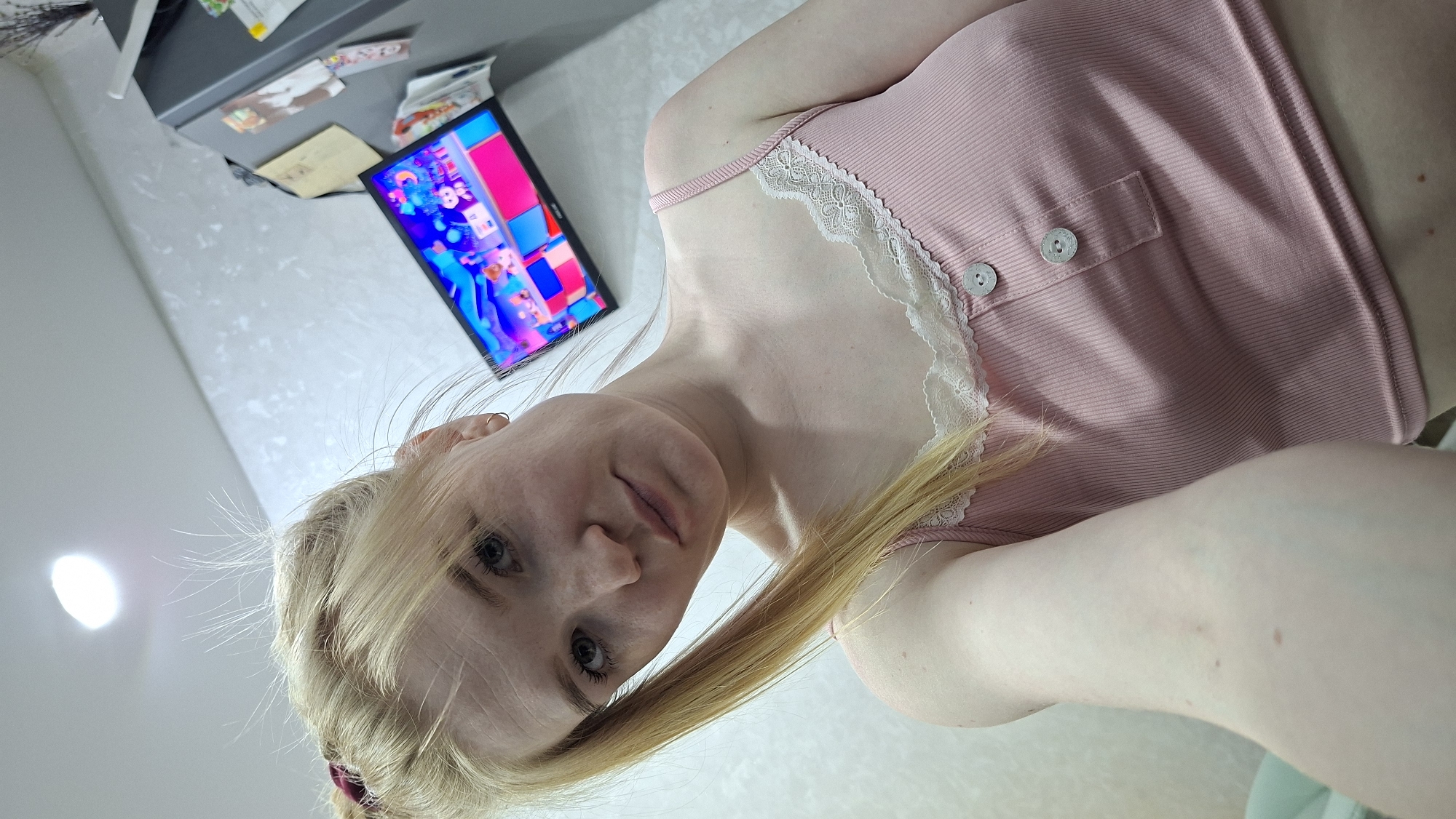 A woman with blonde hair and fair skin is wearing a pink top with lace trim and buttons. She is positioned in front of a wall with a television mounted on it. The television screen displays colorful cartoon imagery. Above the television, there is a cabinet with magnets and other objects attached to it. The location is potentially in an indoor environment, specifically in Chaikovsky, Russia, based on the request.