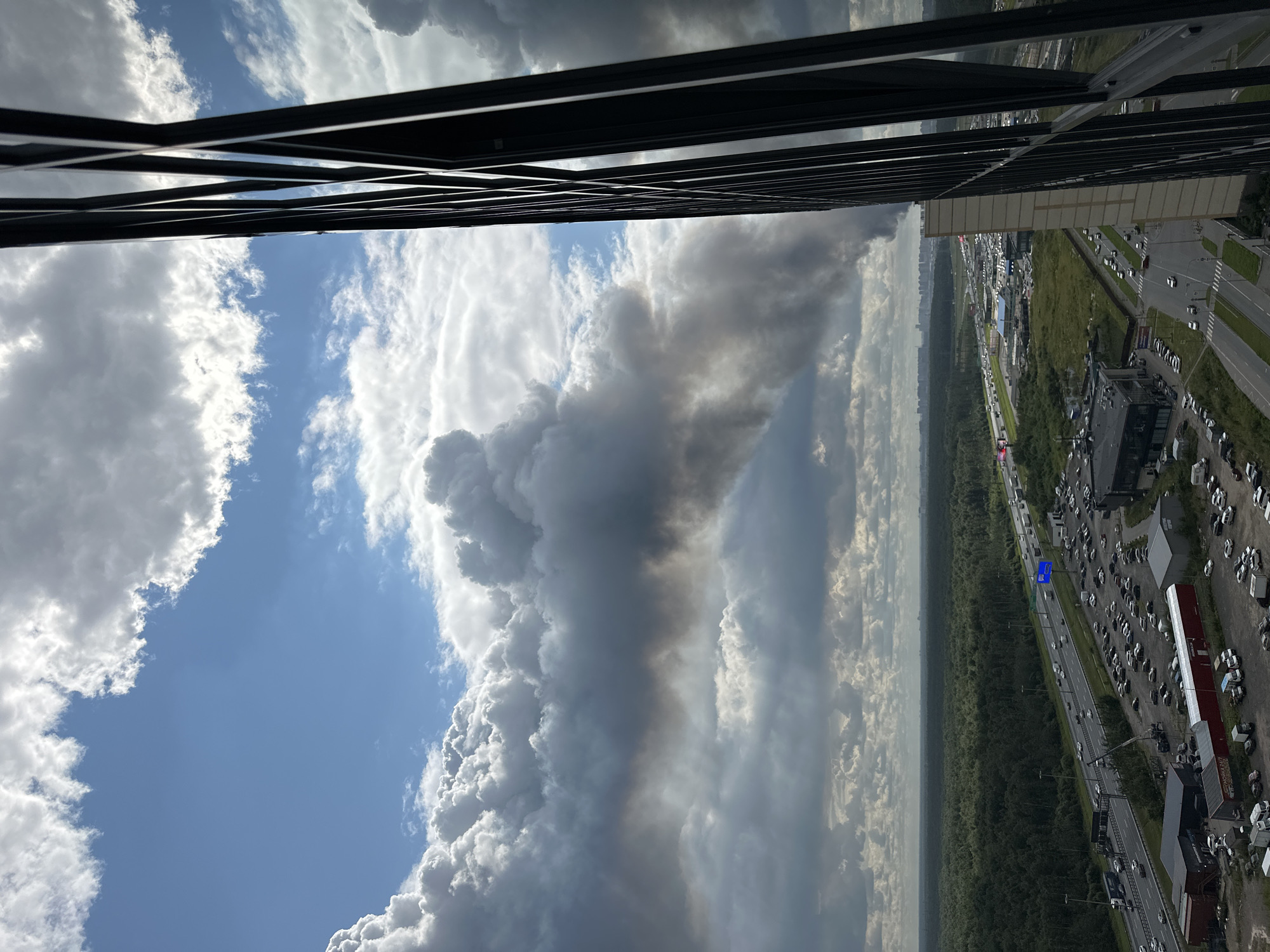 An aerial, off-center view captures a plume of dark smoke rising into a bright blue sky with scattered white clouds, seen from the perspective of a tall building with dark vertical framing in the foreground. The smoke originates from beyond a dense green forest that lines a multi-lane highway filled with moving vehicles. Adjacent to the highway are large parking lots with numerous cars and several industrial-style buildings. The smoke is a significant visual element, extending horizontally across the middle ground of the image. The cityscape is visible in the far distance on the horizon.