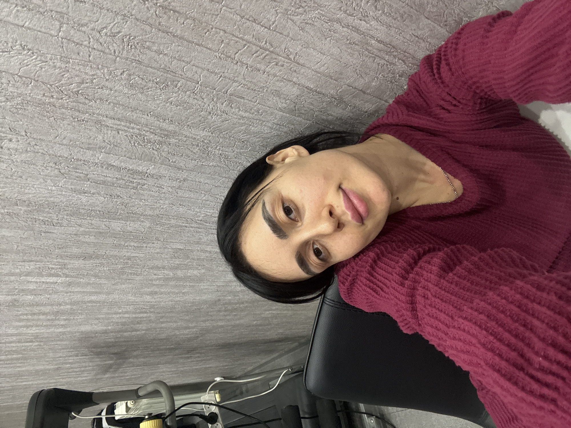 An indoor, chest-up selfie-style portrait features a female individual with dark hair and fair skin. She wears a long-sleeved, deep maroon textured sweater and a thin silver chain necklace. Her head is slightly tilted to the right, and she looks directly at the camera with a neutral expression. She is seated, with the upper back of a black office-style chair visible behind her left shoulder. The background consists of a textured light gray wall with vertical, irregular patterns. To the upper left, a portion of a gray exercise bike, including a handlebar and a yellow adjustment knob, is visible. Below the bike, a white power strip with multiple outlets is mounted on the wall, from which several black and white electrical cords hang. The wall in the immediate upper left corner appears to be a darker gray. The precise location within the city of Tbilisi, Georgia, cannot be determined from the visual evidence present in this image.
