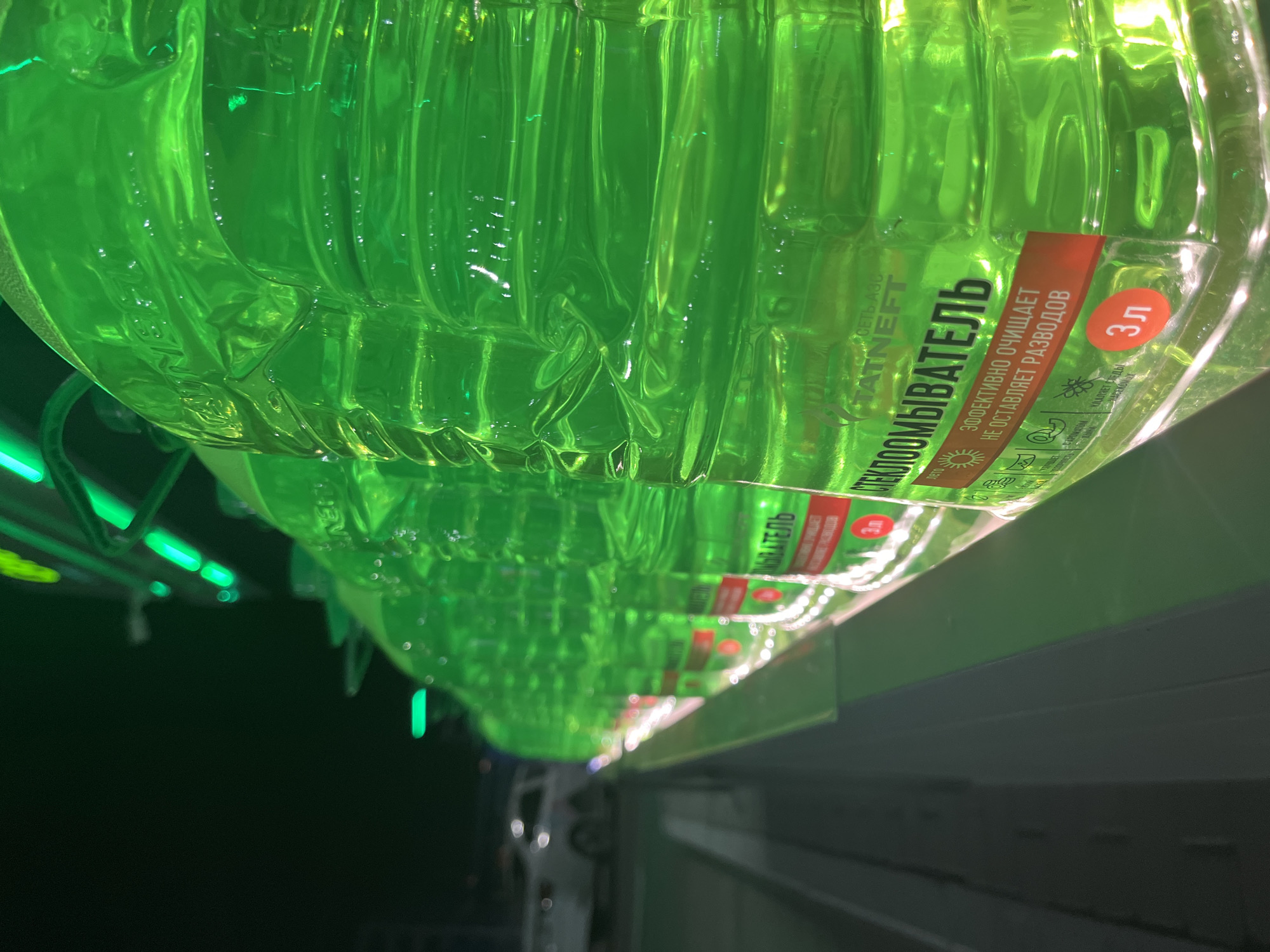 The image shows multiple 3-liter bottles of green windshield washer fluid with red labels, arranged in a row, presumably on a store shelf. The labels prominently feature the Russian word "СТЕКЛООМЫВАТЕЛЬ," which translates to "windshield washer." The brand "TATNEFT" is also visible on the labels. The bottles are illuminated by green lighting, potentially LED strips above the shelf. The scene is likely a retail setting, such as a gas station or auto parts store. While the city is mentioned as Kazan, Russia, there are no specific visual cues in the image itself to confirm this location.