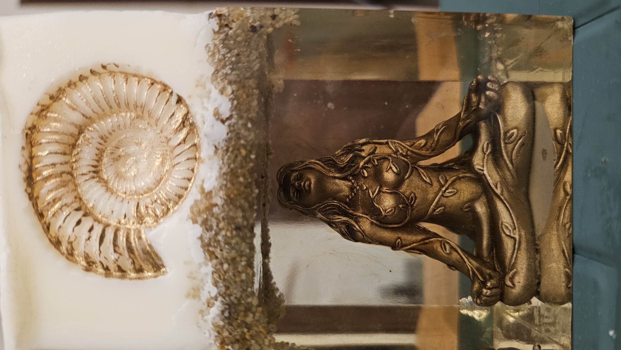 The image shows a close-up of an object with two distinct sections. The upper section is white and features a golden ammonite fossil embedded near the top. Below this, a layer of sand and small pebbles separates it from the lower section. The lower section is made of clear material, possibly resin, and contains a golden figurine of a woman in a meditative pose. The figurine is depicted with long hair and is adorned with vine-like patterns. The background within the clear section is blurred and reflects light, suggesting it is housed in a display. The object rests on a dark teal surface. The location in Baan Chaweng, Thailand is not discernible from the image itself.