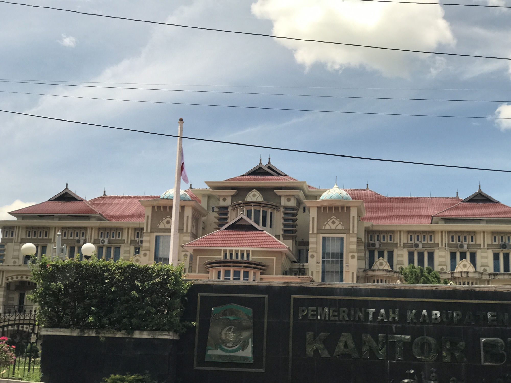 The image shows the Kantor Bupati Aceh Besar, the office of the regent of Aceh Besar, located in Jantho, Indonesia. The building is a large, two-story structure with a red-tiled roof, light-colored walls, and numerous windows. A flag pole with the Indonesian flag stands to the left of the building's main section. In front of the building is a dark wall with a sign that partially obscures the view. The sign has an emblem and the words "PEMERINTAH KABUPATEN" and "KANTOR B..." visible. A green hedge and a black metal fence are visible in the foreground. Electrical wires run across the sky above the building. The sky is blue with some white clouds.