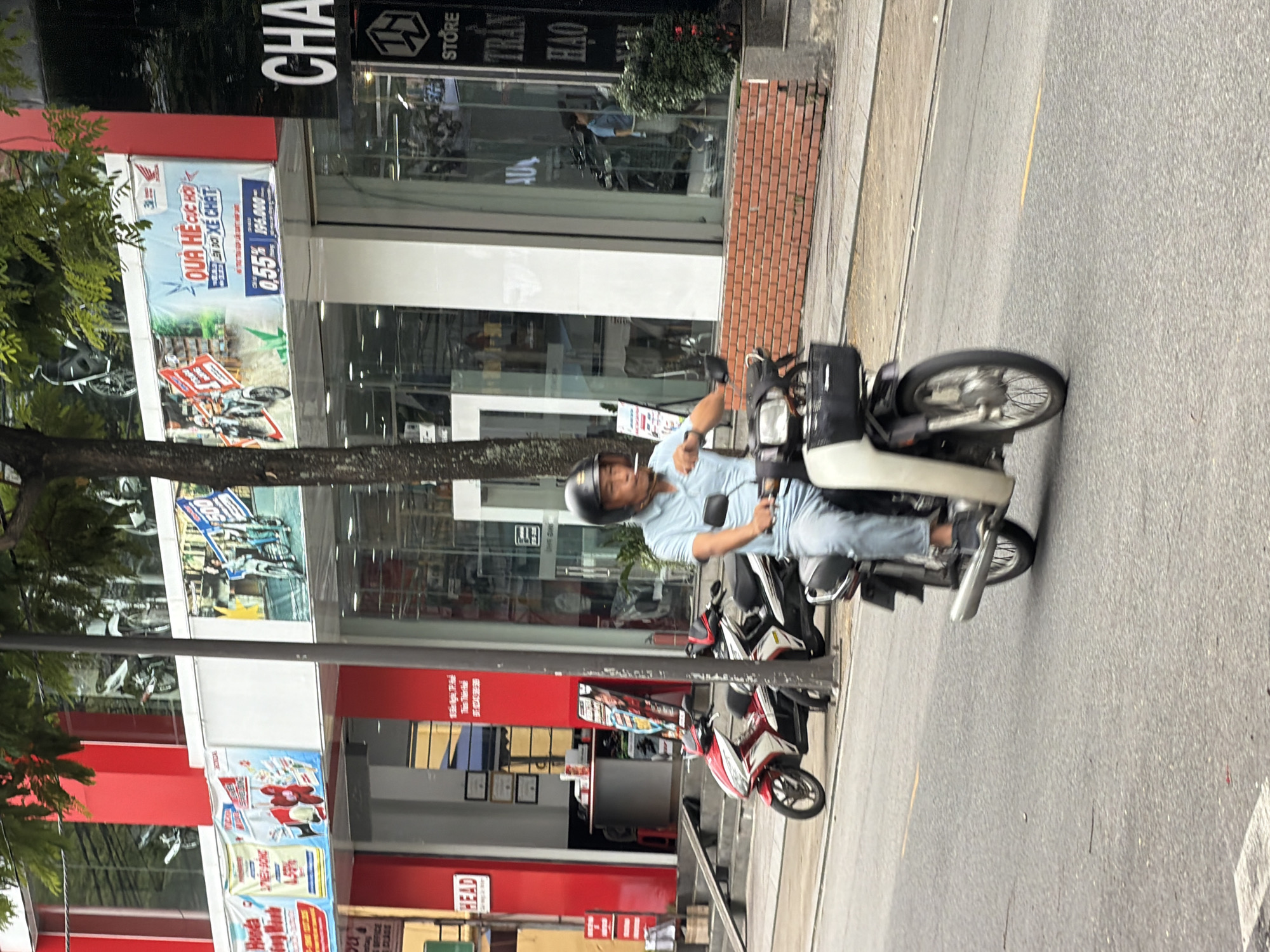 In Thuận Hóa Ward, Vietnam, a man wearing a light blue short-sleeved shirt and light blue pants rides a scooter. He is wearing a black helmet and smoking a cigarette. The scooter is white and black with a black basket attached to the front. In the background, there is a red and white building with large glass windows. Several motorbikes are parked outside the building. A large banner with Vietnamese text and images of motorbikes is displayed on the building's facade. A tree with green leaves is visible in the foreground, partially obscuring the building. The scene takes place on a street with a paved road surface.