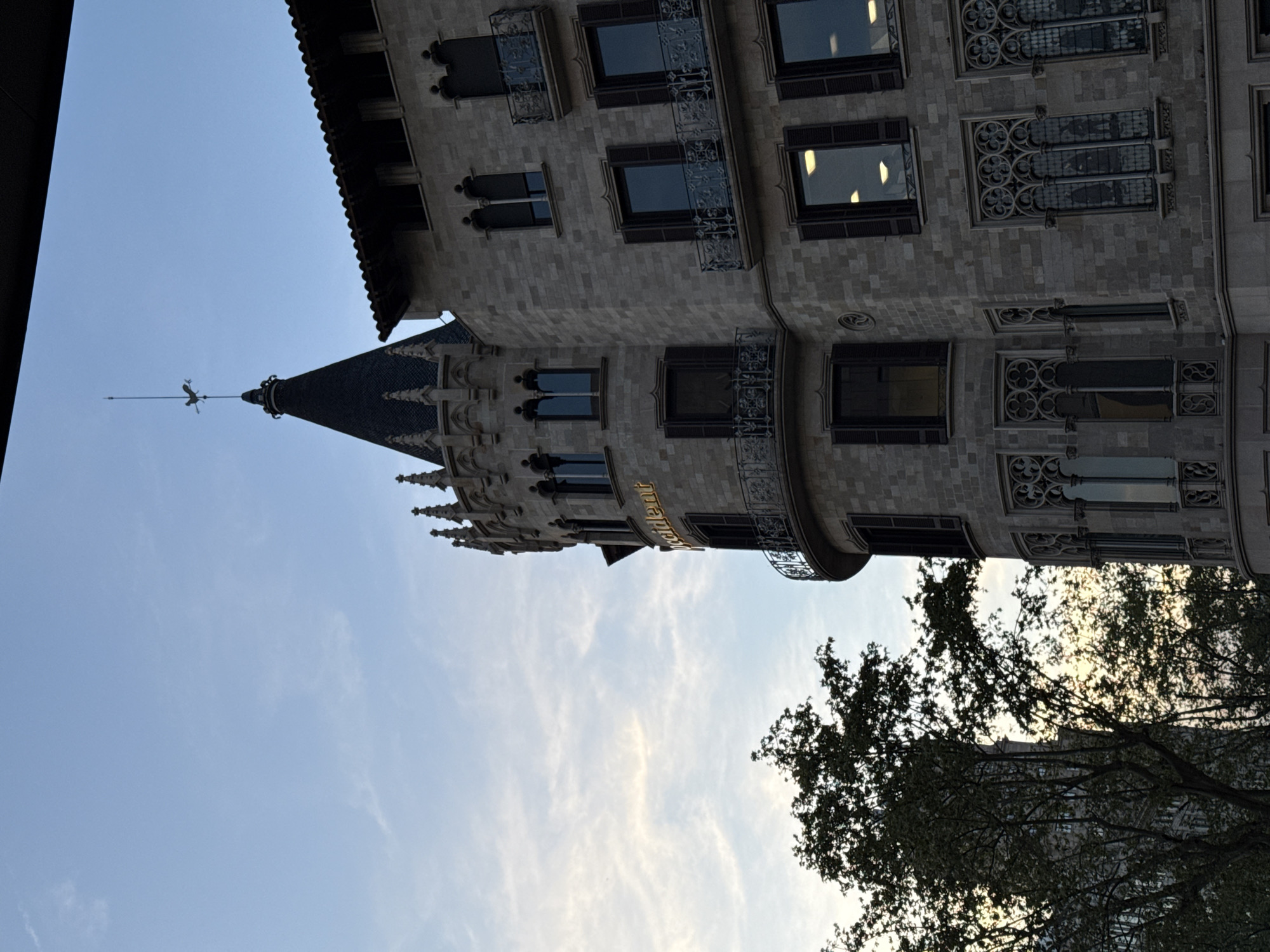 The image depicts the Casa Amatller, a modernist building in Barcelona, Spain, designed by architect Josep Puig i Cadafalch. The building's distinctive turret and ornate facade are visible against a blue sky. Trees are seen in the foreground, and part of a building is visible on the left of the image.