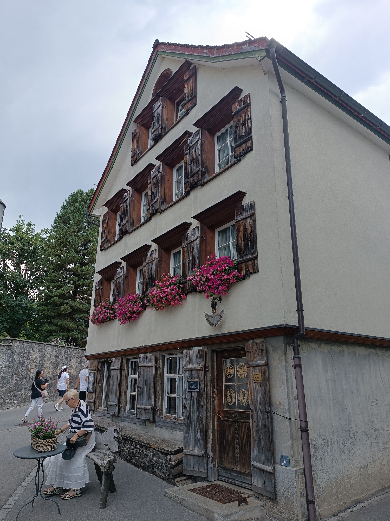 The image shows the exterior of a traditional wooden-shuttered building in the city of Appenzell, Switzerland. The building has several stories with multiple windows, many adorned with vibrant pink flowers in window boxes. A woman in a striped shirt and white skirt is seated on a rustic wooden bench outside the building, tending to a basket of flowers on a small, round table. In the background, other individuals are walking along a cobblestone path, and a stone wall lines the street. The building's facade features rustic wooden doors and windows, and a downspout runs vertically along the side. The overall scene depicts a quaint and charming street view in Appenzell.