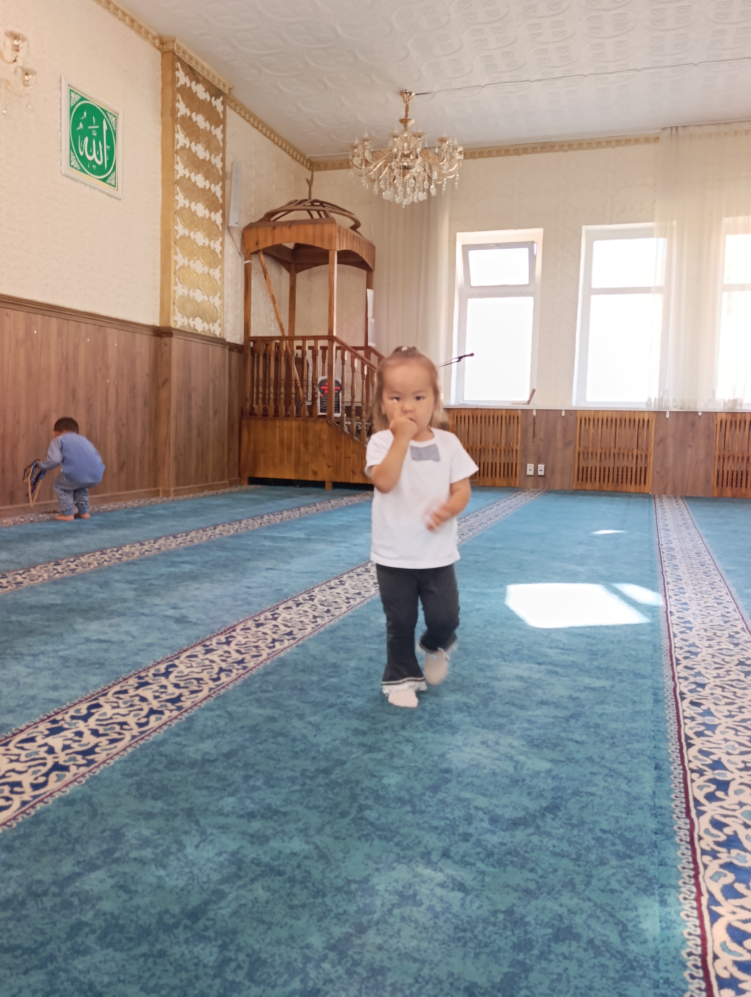 A wide-angle shot captures the interior of a mosque in Naryn City, Kyrgyzstan, characterized by a vast expanse of teal-colored carpet with an intricate blue and white patterned border. In the foreground, a young girl with dark hair tied back, wearing a white t-shirt and dark pants, stands with her right hand near her mouth, looking towards the camera. Behind her, to the left, a younger child in a blue outfit is crouched down, appearing to be interacting with something on the floor near a wooden paneling. The mosque's interior features ornate walls with gold-colored decorative patterns, a wooden minbar or pulpit, and a large chandelier hanging from the textured ceiling. Sunlight streams through tall windows, illuminating the space and casting shadows on the carpet. The overall impression is of a serene religious space with children present.