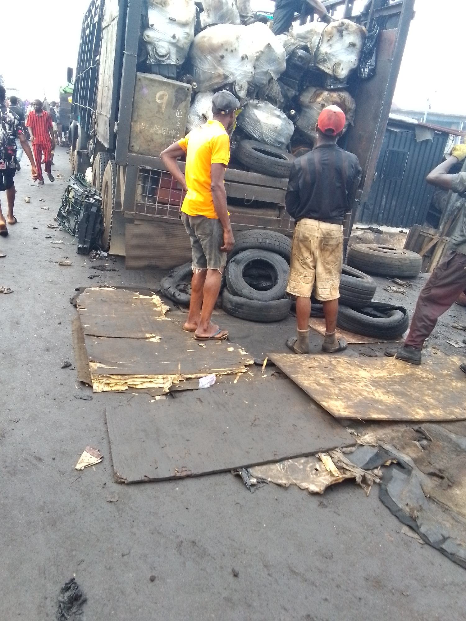 The image captures a scene in Onitsha, Nigeria, likely a roadside or informal waste disposal area. A dump truck is loaded with discarded white plastic containers, tires, and other trash. Several people are present, seemingly involved in unloading or scavenging. One man in an orange shirt stands with his hand on his hip, another in a black shirt and red cap stands nearby. The ground is littered with debris, tires, and wooden planks. There are additional people and traffic visible in the background, suggesting an urban environment.