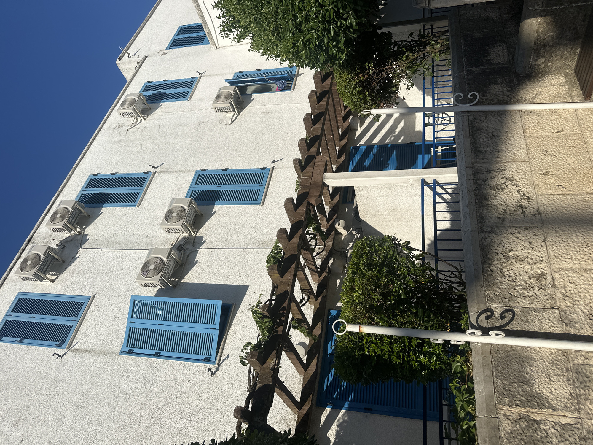 This image shows the exterior of a white, textured building with multiple blue-shuttered windows. Several air conditioning units are mounted on the exterior wall. A wooden pergola with some green foliage extends across the lower part of the building, casting shadows. In the foreground, there is a stone wall and a white pole with decorative metal elements. The scene appears to be in a sunny outdoor environment, with a clear blue sky visible above. The architecture and the presence of air conditioning units suggest a warm climate, possibly a tourist area. Without further information, the specific location within Budva, Montenegro, cannot be determined solely from the visual elements.