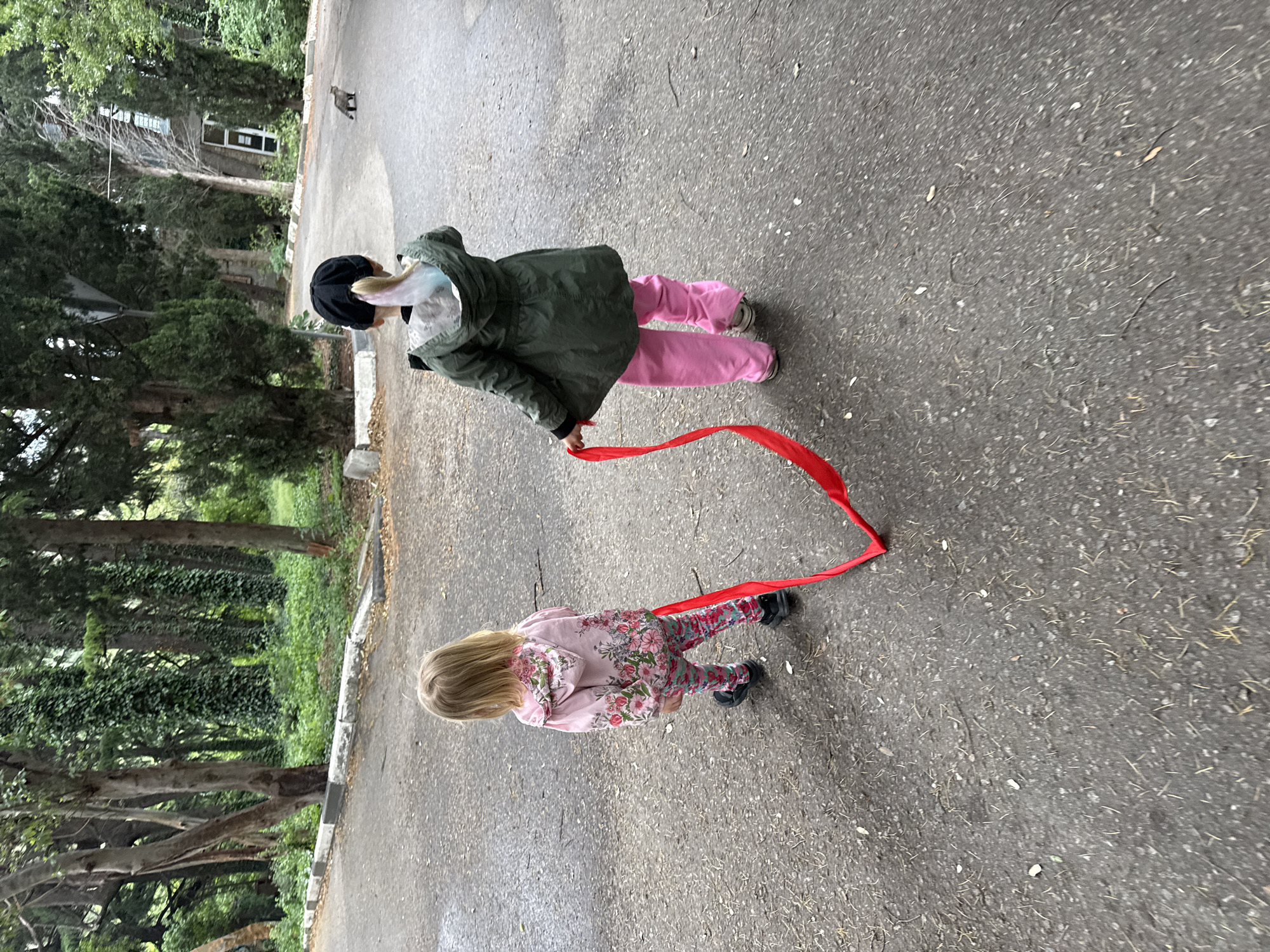 Two children are walking along a paved surface in an outdoor environment. The child in the foreground is wearing a light-colored floral patterned outfit. The child in the background is wearing an olive green jacket, pink pants, and a black hat. A red ribbon connects the children. Trees and vegetation line the side of the pavement. A building is visible in the background. An animal, possibly a cat, is observed in the distance.