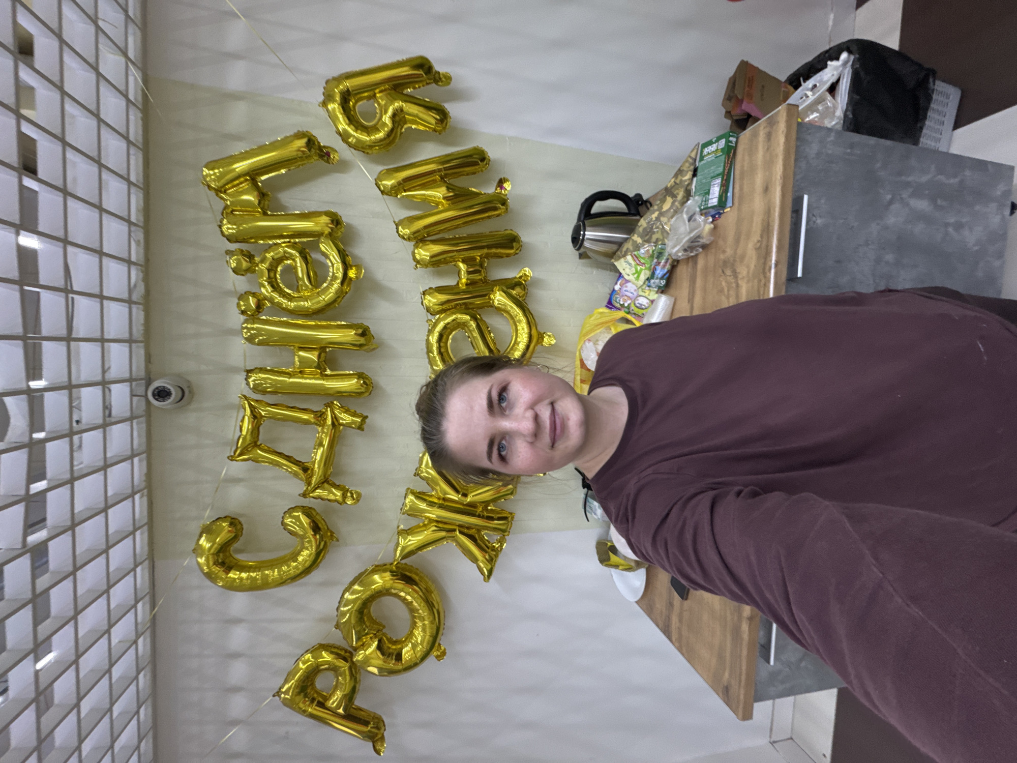 A young woman poses for a photo in a room with gold foil balloons spelling out a message in Russian. The location is possibly in Chaikovsky, Russia, considering the text on the balloons. The room appears to be a kitchen or dining area, with a countertop, a microwave, and other kitchen items visible in the background. The woman is wearing a maroon-colored sweater. The balloons are attached to the wall, with a security camera also visible in the upper left corner.