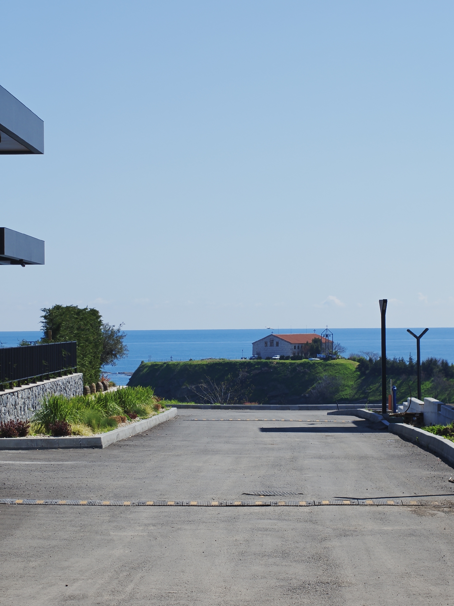 The image depicts an outdoor scene with a paved parking area in the foreground. The parking area has speed bumps running across it. On either side of the parking area are landscaped areas with various plants and greenery. In the midground, there is a grassy cliff overlooking the sea. A building with a red-tiled roof stands on the cliff. In the background, the blue sea meets a clear blue sky. The left side of the image shows parts of a modern building with a dark-colored exterior. Based on the architectural style and environment, the location is possibly Tsarevo, Bulgaria. There are no people visible in the image. Street lamps are placed on the right edge of the cliff. A stone wall is located behind the left side of the parking area.