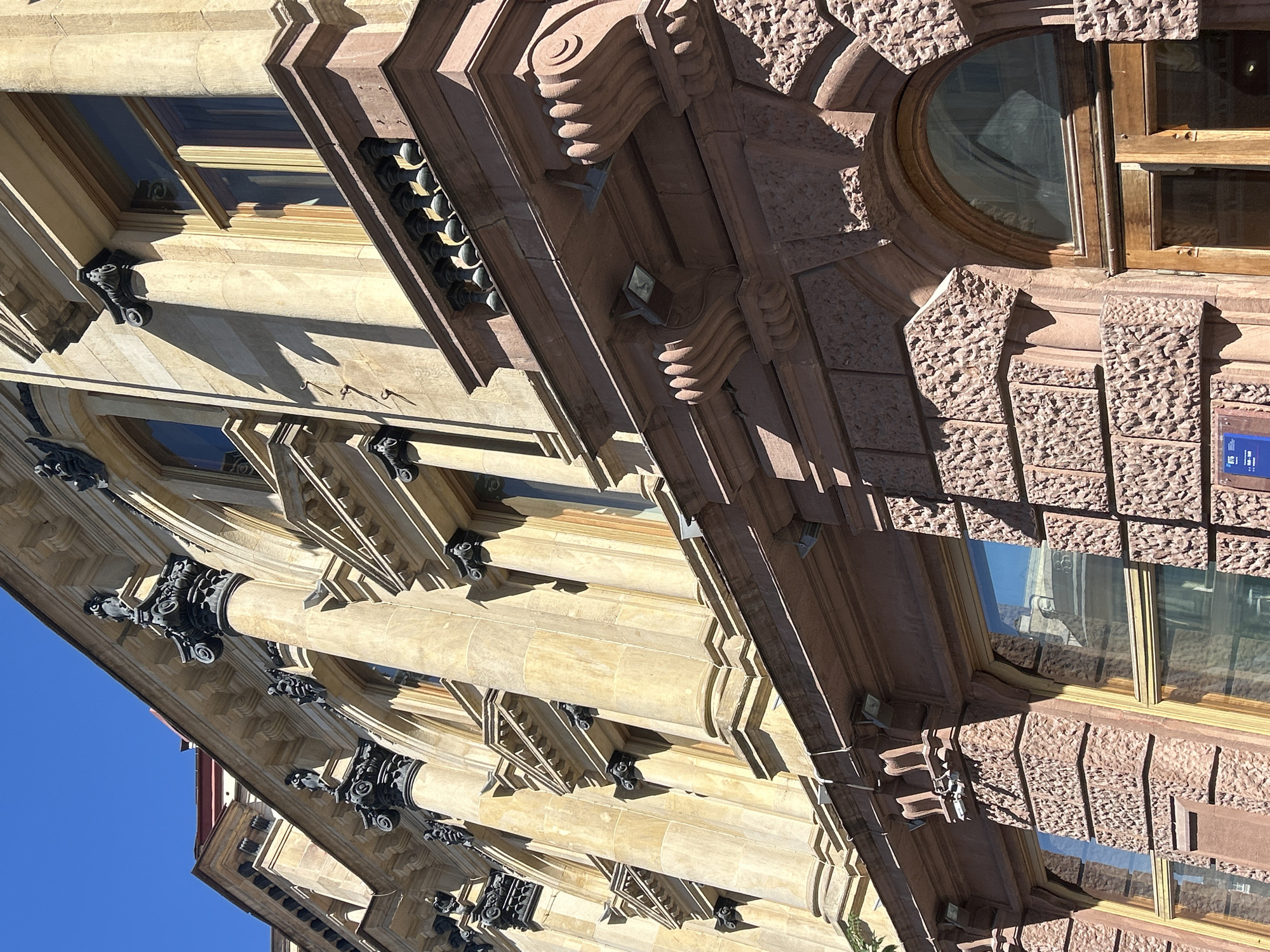 The image shows the facade of an ornate building with a light blue sky in the background. The building's architecture features columns with decorative capitals, arched windows, and a balcony with a balustrade. The stonework is in two distinct colors, a lighter beige on the upper floors and a darker reddish-brown on the lower floors. The lower section of the building has a rough, textured stone finish, and there are large wooden doors at the bottom. The sunlight casts strong shadows, highlighting the architectural details. Several small light fixtures are mounted on the facade above the lower level.