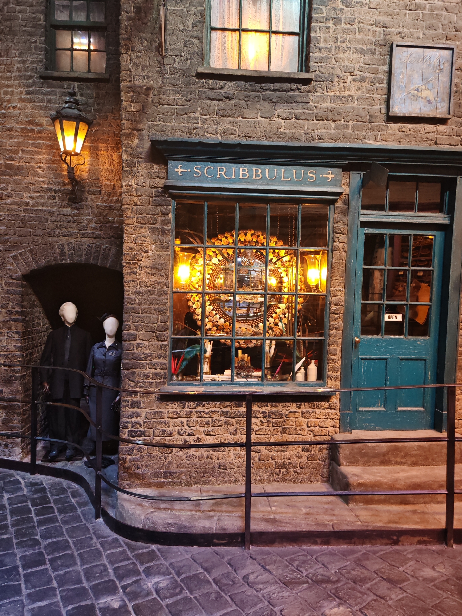 The image captures a street scene within the Warner Bros. Studio Tour London in Watford, United Kingdom. The scene recreates a segment of Diagon Alley from the Harry Potter series.
A weathered, brick facade dominates the view. Above the window of the establishment "Scribbulus" is written. To the left, a stone archway houses two mannequins dressed in dark clothing. One is a male figure in a suit, and the other appears to be a female figure in a dress and hat. A wrought-iron railing separates the mannequins from the cobblestone street.
To the right of the archway, the store window is filled with an elaborate display, illuminated by warm, bright lights. The reflections of the glass obscure the view inside, although a decorative mirror surrounded by shells is partially visible. Adjacent to the window is a turquoise door marked with an "OPEN" sign. The cobblestone street extends into the foreground, leading to a set of steps that lead up to the door.
A gas-style lantern illuminates the street, affixed to the brick wall above the archway. The overall atmosphere evokes a sense of historical charm and mystery.