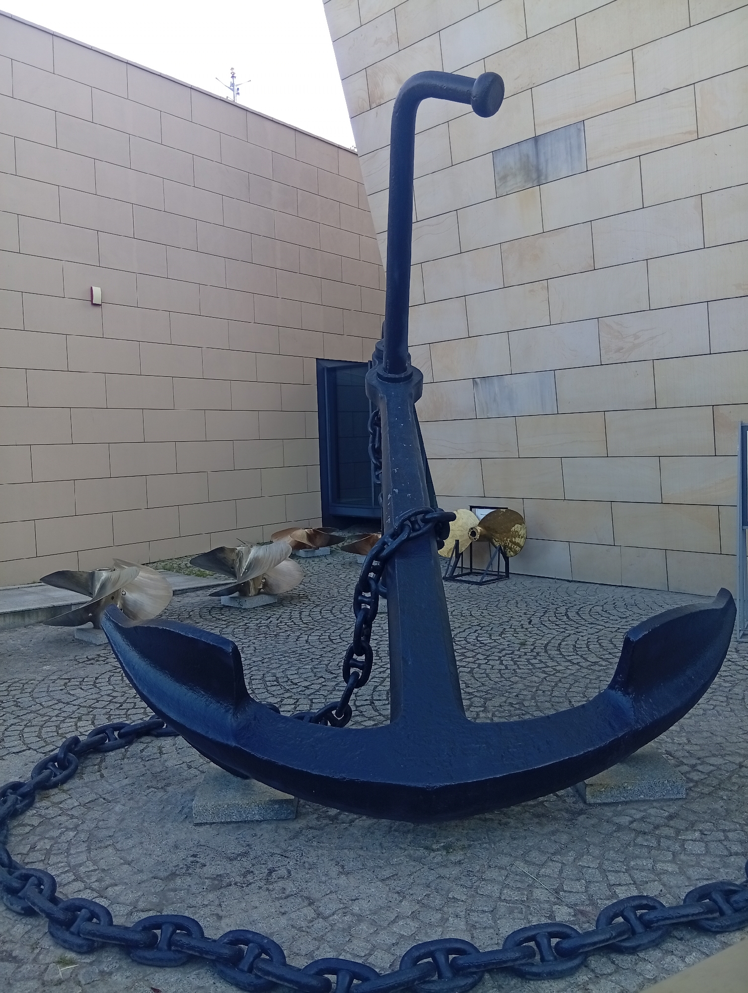 An imposing, dark blue anchor with a thick chain lies on a cobblestone surface in front of a beige brick building. The anchor is positioned in the foreground, dominating the lower half of the image, with its arms spread wide. The building's facade is made of rectangular beige bricks, laid in a horizontal pattern. Behind the anchor, two large, brass-colored ship propellers are displayed on stands. One propeller is larger and more prominent, while another is slightly behind and to its left. A dark blue door is visible within an alcove of the building. The scene appears to be an outdoor exhibition, likely part of the National Maritime Museum in Gdynia, Poland, given the maritime artifacts. The lighting suggests it is daytime, with soft, diffused light.