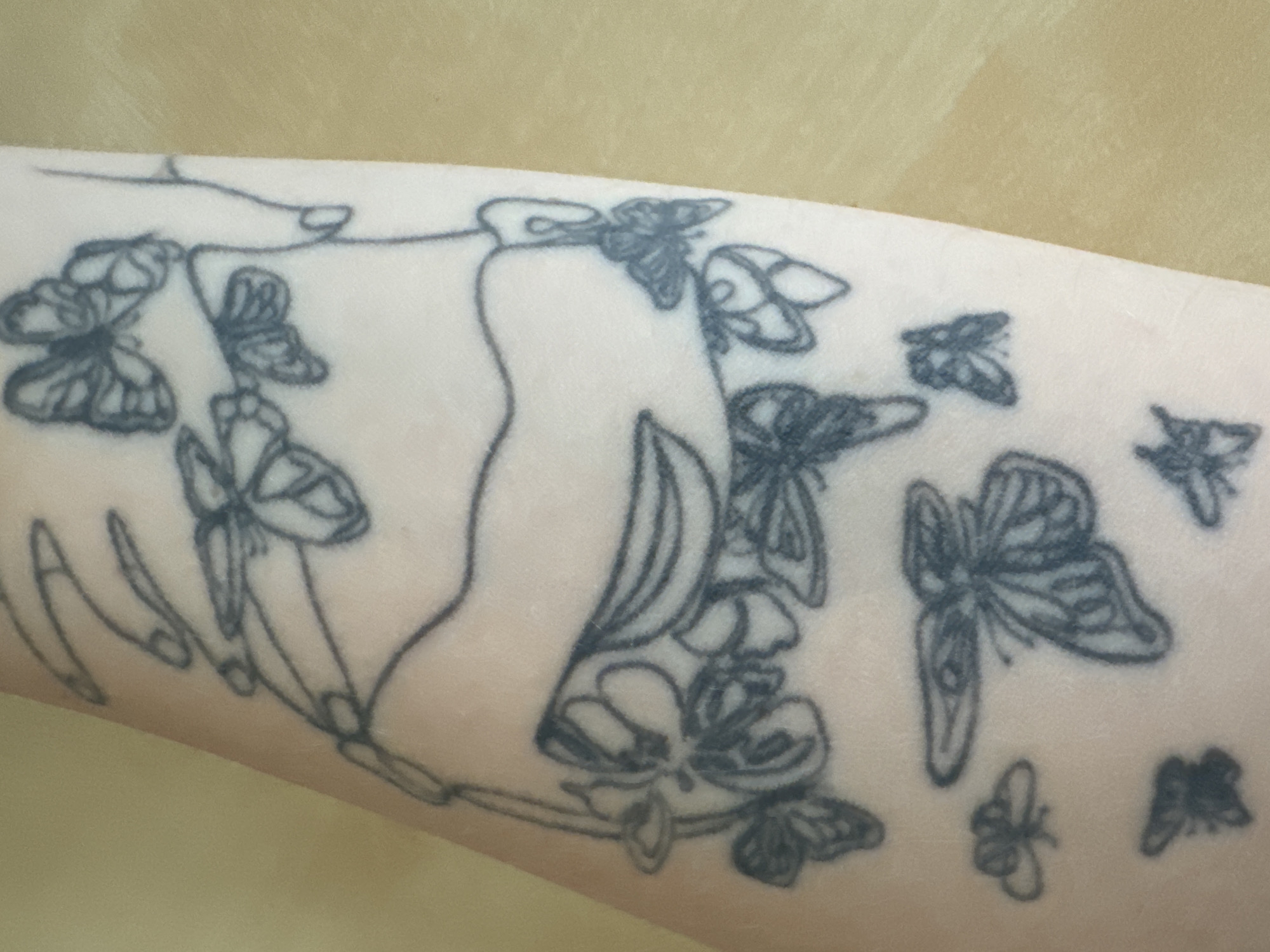 This is a close-up, eye-level, full-frame shot of a tattoo on a person's arm. The tattoo depicts a branch with several butterflies and what appears to be a stylized hand or flower. The tattoo is rendered in black ink and appears to be in progress or faded. The skin tone is fair. The background is a plain, light yellow surface, possibly a table. The image is not in Murcia, Spain.