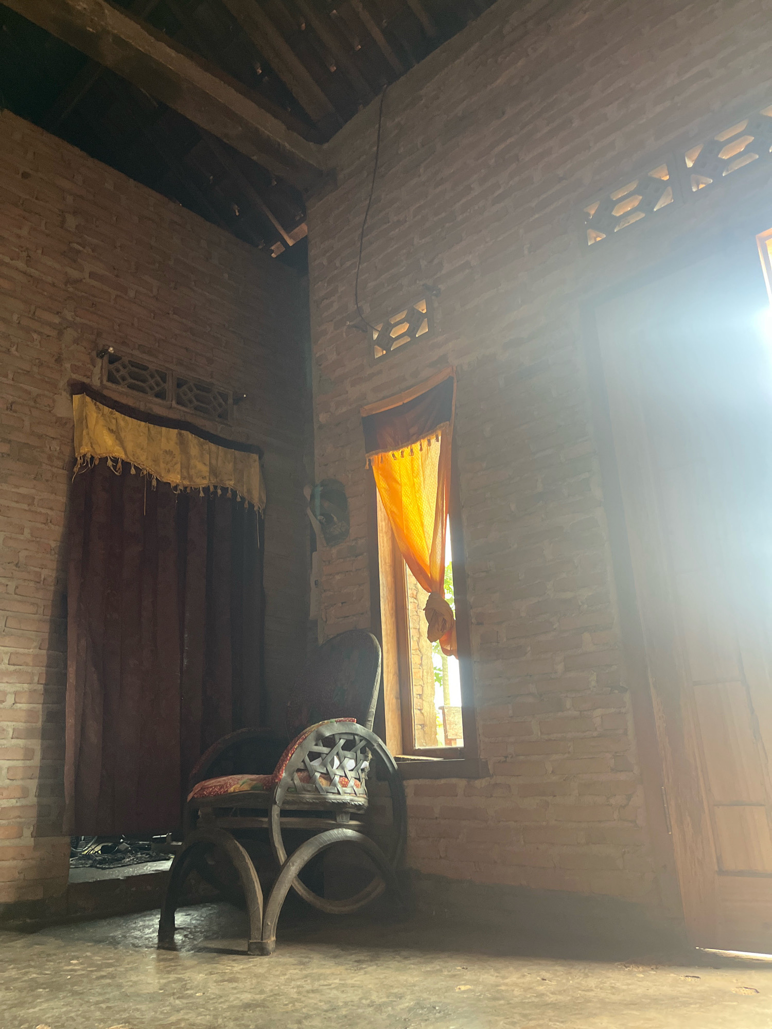 This is a medium shot, eye-level, interior photograph of a rustic room, possibly in Sidomulyo, Indonesia. The room is constructed of unpainted brick, with visible wooden beams supporting the roof. Sunlight streams in from a window on the right, illuminating dust motes in the air and casting a strong glare.
In the center of the frame, a dark-colored wooden armchair with a intricately carved backrest and armrests is positioned on the concrete floor. The seat of the chair appears to be upholstered with a patterned fabric. To the left of the armchair, a tall, dark brown curtain is drawn, with a decorative yellow fabric trim at the top. Above the curtain, a small louvered vent is visible in the brick wall.
On the right side of the room, another window is draped with a vibrant orange curtain, which is partially open, allowing sunlight to flood the space. To the right of this window, a wooden door is partially visible. The floor is concrete and appears worn. The overall impression is one of simple, traditional living, with natural light playing a significant role in the atmosphere. There are no people or animals present in the image.