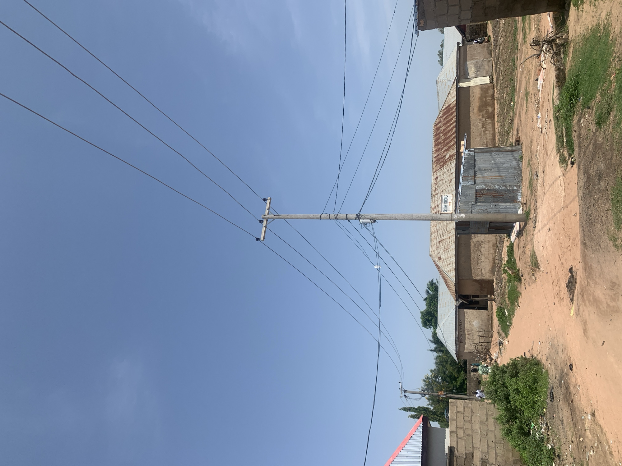 This is a medium-distance, eye-level, outdoor photograph taken in Jauro Yinu, Nigeria, under a clear blue sky with a few wispy clouds. The scene depicts a dirt road winding through a residential area with several buildings. A prominent wooden utility pole with multiple power lines stretches across the upper portion of the image.
The buildings are constructed with mud or concrete block walls and corrugated metal roofs, some of which show signs of rust. To the left, a building with a red-trimmed metal roof is visible, adjacent to a brick wall with overgrown green bushes and debris. In the center, a small, corrugated metal structure stands in front of a building labeled "DESTINY SCHOOL" with an arrow pointing to the right. Further down the road, other buildings are visible, some with their roofs partially obscured by trees.
There are at least two individuals visible: one man wearing a white shirt and dark pants walking on the left side of the road, and another person in a green and white checkered shirt walking further down the road. The ground is mostly bare red dirt with patches of dry grass and scattered debris. The environment appears to be a rural or semi-urban area with a focus on basic infrastructure and housing.