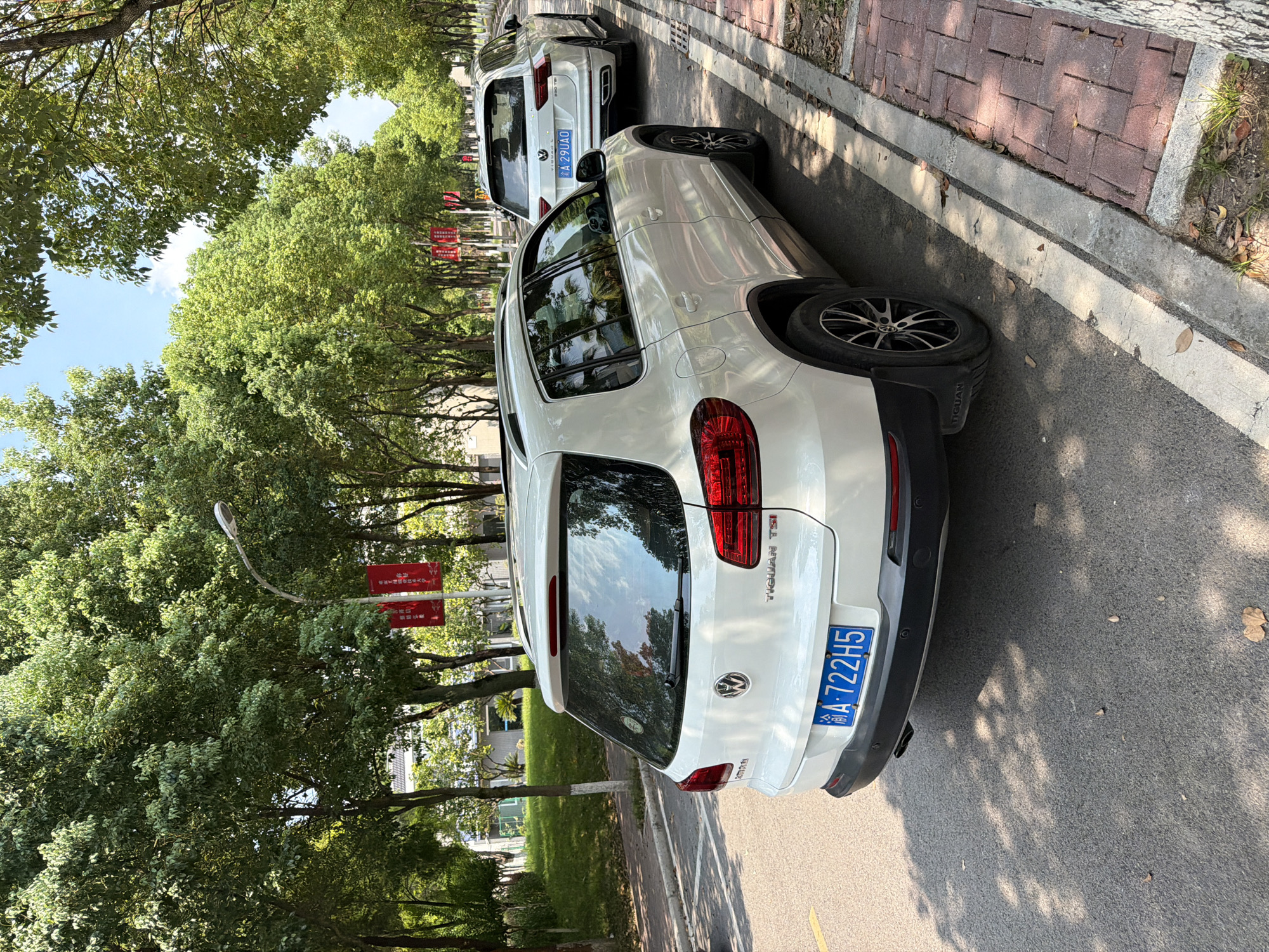 The image depicts a street scene in Jiangjin, China, lined with lush green trees casting dappled shadows on the asphalt. Two white Volkswagen Tiguan SUVs are parked along the right side of the road. The foreground car, a Tiguan TSI, is white with a blue license plate reading "A-722H5". Its rear bumper has a subtle black diffuser and two exhaust pipes. The second Tiguan, further down the street, is also white and displays a license plate "A 29UAO". Red banners are visible hanging from lampposts between the trees, suggesting a festive or official occasion. The ground beside the road is paved with red bricks, bordered by a concrete curb, and some dry leaves are scattered on the pavement and the brick walkway. The sky is bright blue with a few scattered clouds, indicating a sunny day. The overall atmosphere is calm and serene, with the cars and trees forming a typical urban streetscape.