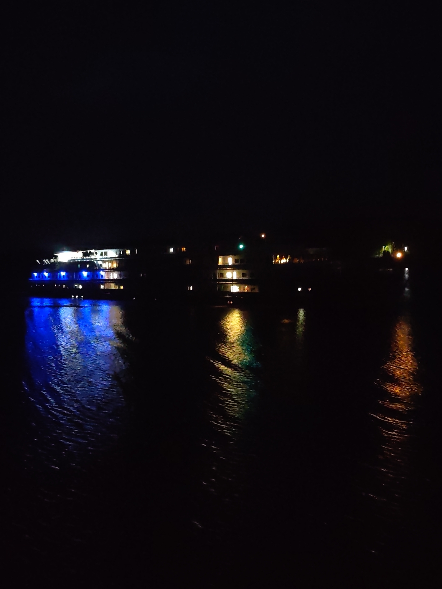 The image depicts a night scene on the water in Basharino, Russia. A multi-decked boat is illuminated with blue lights on its front section. Other buildings are visible in the background with yellow and green lights. The reflections of these lights are visible on the water. The water surface shows ripples and movement. The sky is dark, suggesting a lack of artificial light pollution in the immediate vicinity of the observation point. No people are visible.