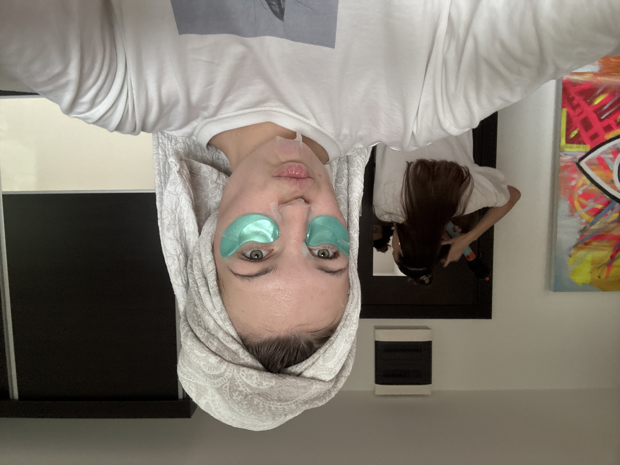 A selfie shows a woman's face upside down. She has a white towel wrapped around her head and green eye patches under her eyes. A clear patch is visible above her lips. She wears a white t-shirt. Reflected in a mirror behind her, a person with long dark hair is visible, bending over. A painting is to the right of the mirror and a black and white cabinet is to the left. A small white appliance sits on a shelf between the mirror and cabinet. The location is likely indoors, in a bathroom or bedroom. The city of Vozdovac Urban Municipality, Serbia is not evident in the image.