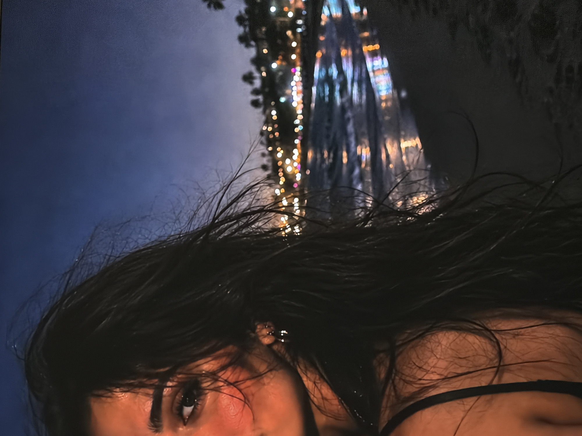 The image presents a close-up, angled portrait of a person, likely a woman, with long, dark hair dominating the foreground. Her head and upper torso are positioned towards the bottom and right of the frame. Her dark hair appears sleek, possibly wet, and falls across her face and body. One dark eye, with a visible pupil and eyelashes, is clearly visible on the left side of her face, gazing towards the viewer. Her skin has a warm undertone, with a soft, reddish hue on her cheek. A small, silver-colored stud earring is discernible in her ear, and a thin, dark strap of clothing, likely from a top or bra, is visible across her chest.
The background is deeply blurred and out of focus, suggesting a nighttime setting. A dark blue-grey sky occupies the upper left quadrant. To the right, numerous small, colorful, out-of-focus lights, resembling fairy lights or holiday decorations, create a bokeh effect. Behind these lights, a larger, vertically stretched, brightly illuminated and blurred structure, possibly a building or an elaborate light display, adds to the festive atmosphere. Sparse, dark foliage is faintly visible, contributing to the depth of field. The overall scene evokes a moody and atmospheric ambiance. No text is visible in the image. The location context is Sofiivska Borshchahivka, Ukraine.