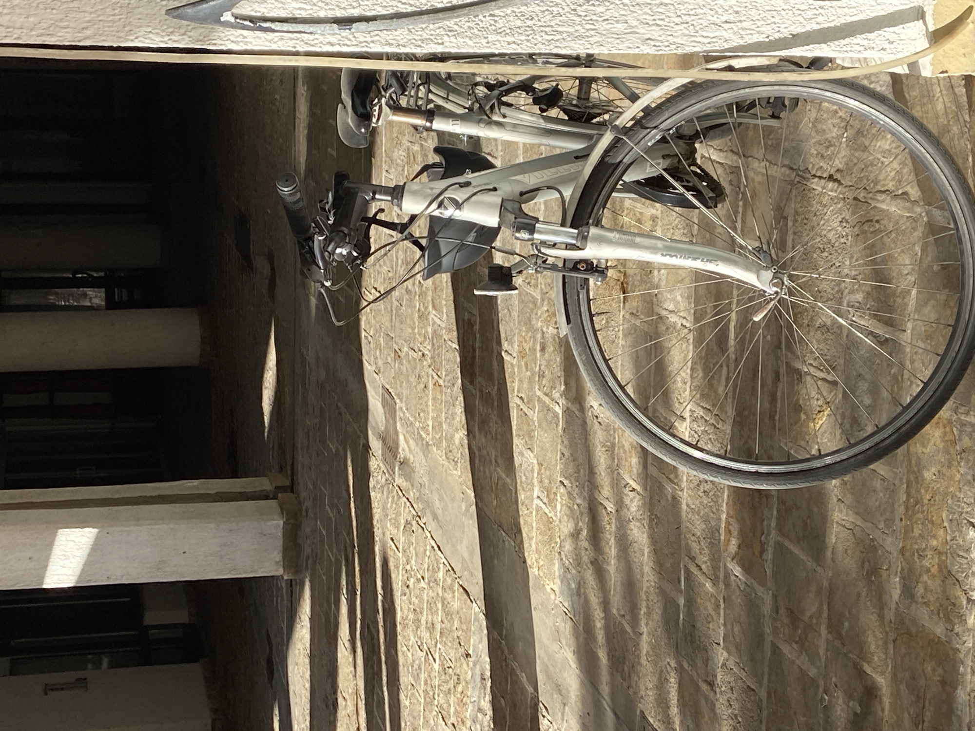 A silver-framed bicycle with road tires leans against a stone wall. The wheel spokes and frame are visible in detail. The handlebars and gears are also visible. The background includes an adjacent building with windows and architectural details. The location is Budva, Montenegro.