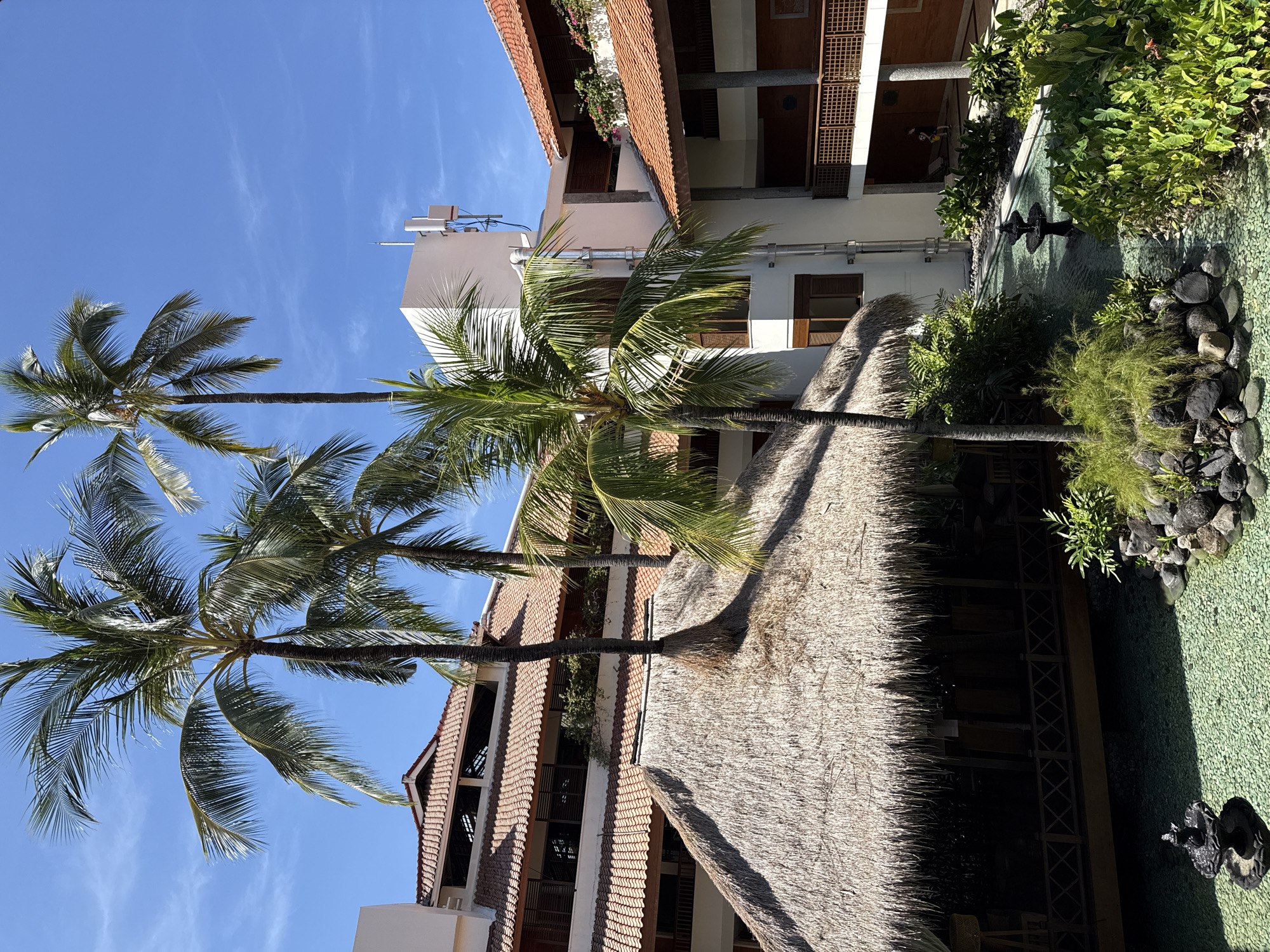 The image shows an outdoor scene in Kuta Selatan, Indonesia, featuring a building complex with a thatched roof extending over an open area. Two tall palm trees are visible against a clear blue sky. The buildings have red tile roofs and multiple stories with balconies or terraces. A water feature or pond with decorative stones and landscaping is present. The scene appears to be a resort or hotel setting. No people are clearly visible in the shot.
