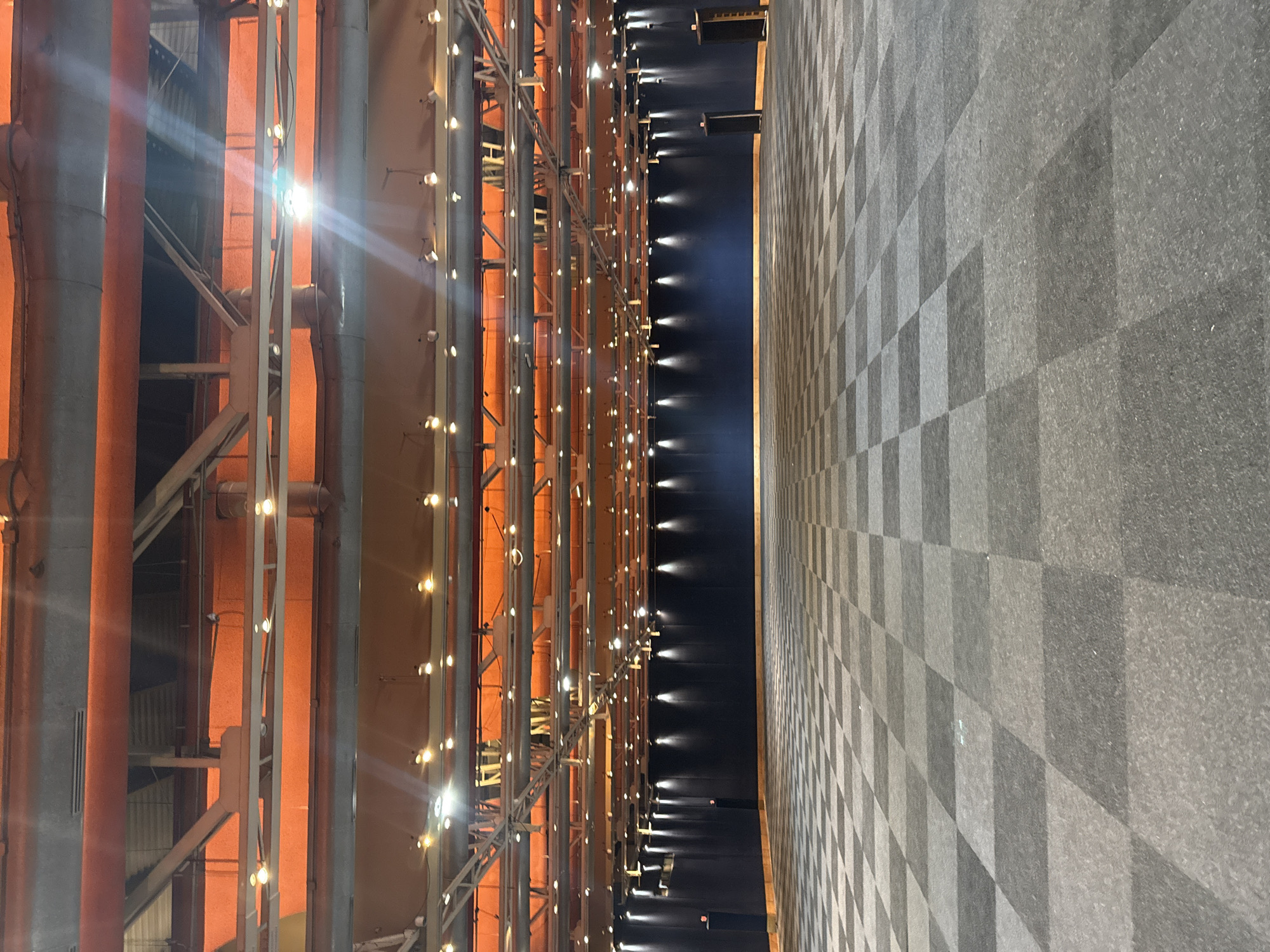 The image displays an architectural interior, presented in portrait orientation, featuring a striking interplay of light, industrial elements, and geometric patterns. On the left, a tall, multi-layered structure is visible, comprising numerous gray vertical pipes or columns and horizontal beams, set against a warm, orange-hued background. This intricate framework is densely adorned with numerous small, bright lights that emit intense glows and prominent lens flares, illuminating the structure and the orange wall. Exposed conduits and wires are visible among the pipes and beams.
To the right, a large floor extends, covered with a distinct checkerboard pattern in varying shades of gray, creating an impression of depth and perspective. A narrow, dark channel or pathway separates the illuminated structure from the patterned floor. This channel also features a series of smaller, subtle lights along its left edge, casting soft reflections onto its dark surface.
The scene is devoid of people and discernible activity, presenting a static, artistic, or industrial-inspired vista. The artificial illumination suggests an indoor setting or a nighttime view. The design elements, including exposed pipes and metalwork, contribute to an industrial yet refined aesthetic. No visible text is present. The context of Puebla, Mexico, suggests this could be a modern art installation, a public building's interior, or a creatively repurposed industrial space.