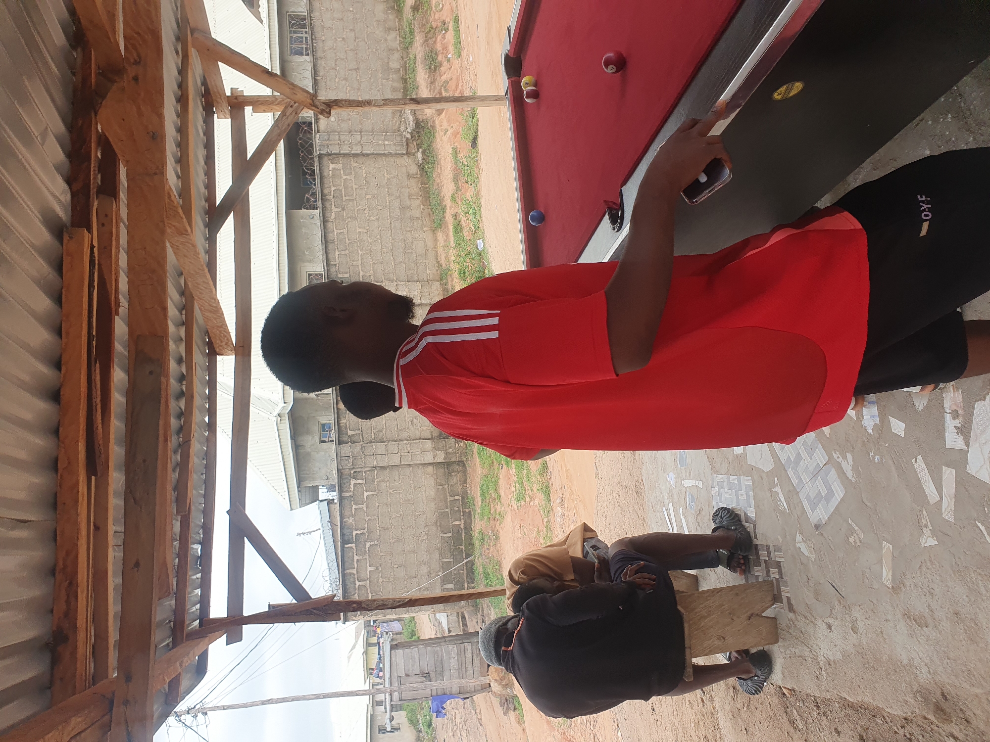 In Lokoja, Nigeria, under a corrugated metal roof supported by rough wooden beams, three individuals are present. One person, facing away from the camera and wearing a red t-shirt with white stripes on the sleeves and black shorts, stands near a red pool table with several balls scattered on it. Another person, also facing away from the camera, is seated on a wooden stool, wearing a black jacket and dark shorts, and appears to be looking at a mobile phone. A third person is seated next to them, wearing a tan-colored shirt and dark shorts, also looking down. The environment appears to be an outdoor recreational area with a dirt ground, scattered debris, and a brick wall in the background. The sky is overcast.