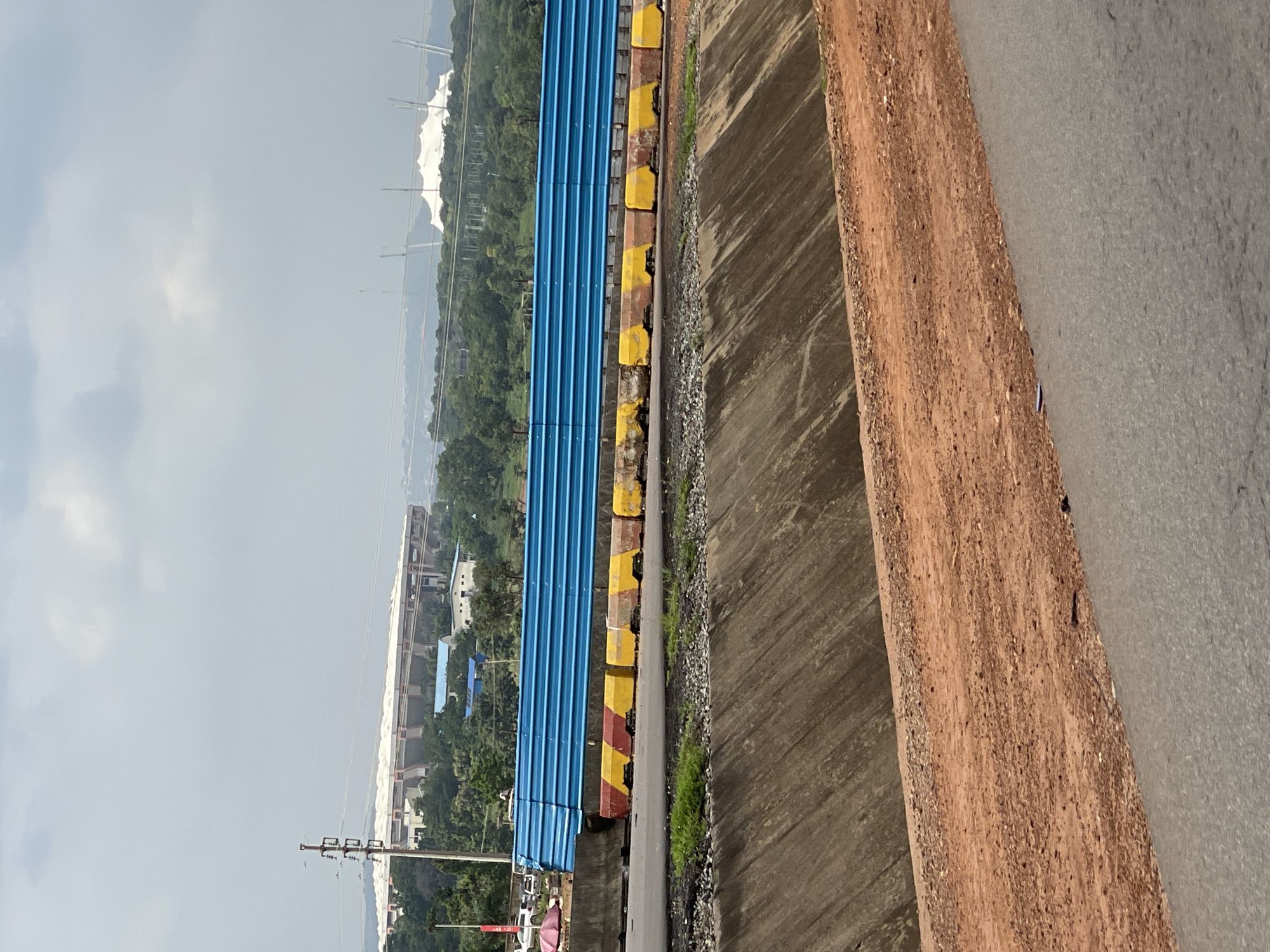 The image depicts a roadside construction scene with a large stadium visible in the background. The stadium, with its white canopy-like roof and distinct architectural features, is likely the Moshood Abiola National Stadium in Abuja, Nigeria, given its prominence and the surrounding landscape. In the foreground, a blue metal barrier with yellow and red concrete barriers underneath separates the road from a ditch. The ditch is lined with concrete and has a slope covered in dirt and gravel. A paved road is visible in the lower part of the image. The sky is overcast with clouds.