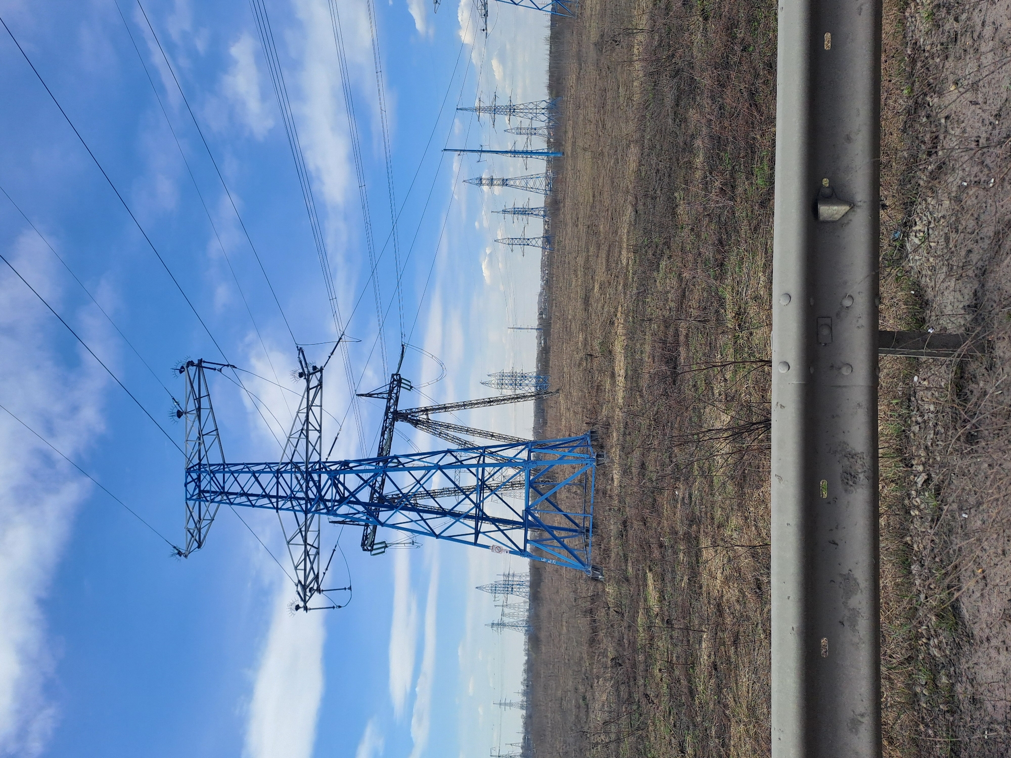 The image depicts a row of high-voltage power line towers extending into the distance under a blue sky with scattered clouds. The nearest tower is painted blue. The ground is covered with dry grass and sparse vegetation. A gray metal guardrail is visible on the right side of the image. The location is near the city of Belaya, Russia. There are no people visible.