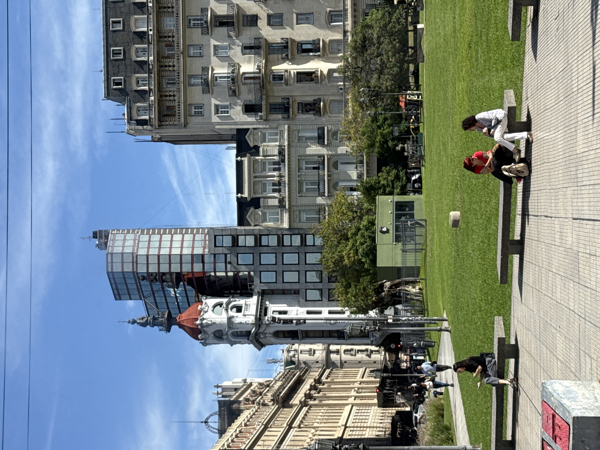 The image depicts a sunny day in Buenos Aires, Argentina, with a view of the Plaza de Mayo and surrounding buildings. The plaza features green lawn areas and paved walkways with benches where people are sitting and relaxing. In the background, there are various architectural styles, including historic buildings with ornate facades, and a modern glass-fronted skyscraper. The sky is clear and blue with a few wispy clouds. The foreground shows benches where people are interacting; for example, one woman in red is sitting next to another in white who leans towards her. There are streetlamps.