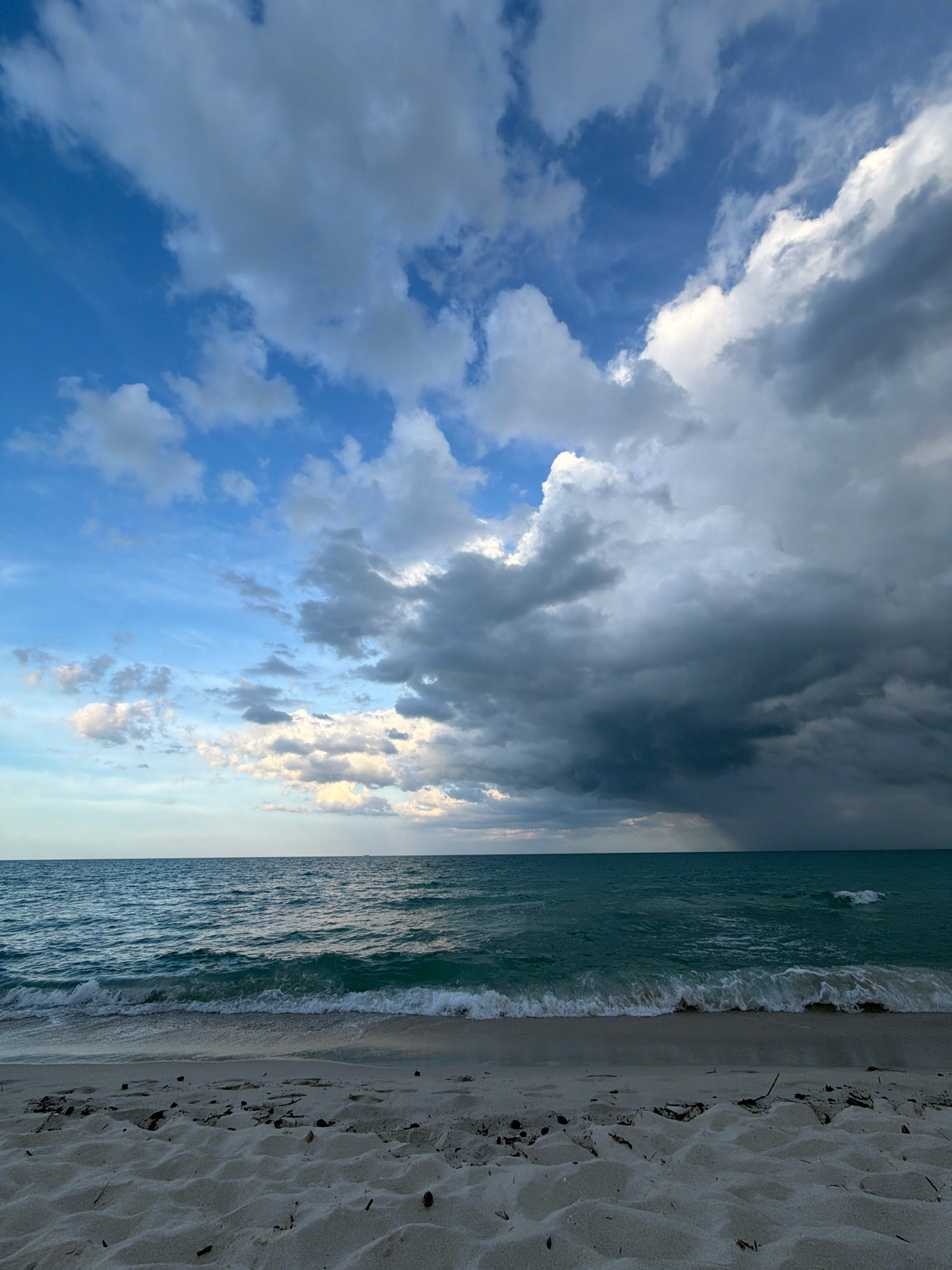 This is a photograph of a beach on the island of Ko Samui in Thailand, taken from a low angle facing the ocean. The sand in the foreground is light-colored and textured with small debris. The ocean is a deep turquoise with gentle waves breaking on the shore. The horizon line is sharp, separating the sea from the sky. The sky is dramatic, filled with a mix of dark, ominous storm clouds on the right and lighter, scattered clouds against a blue sky on the left. The lighting suggests either early morning or late afternoon. There are no people or man-made structures visible in the image.