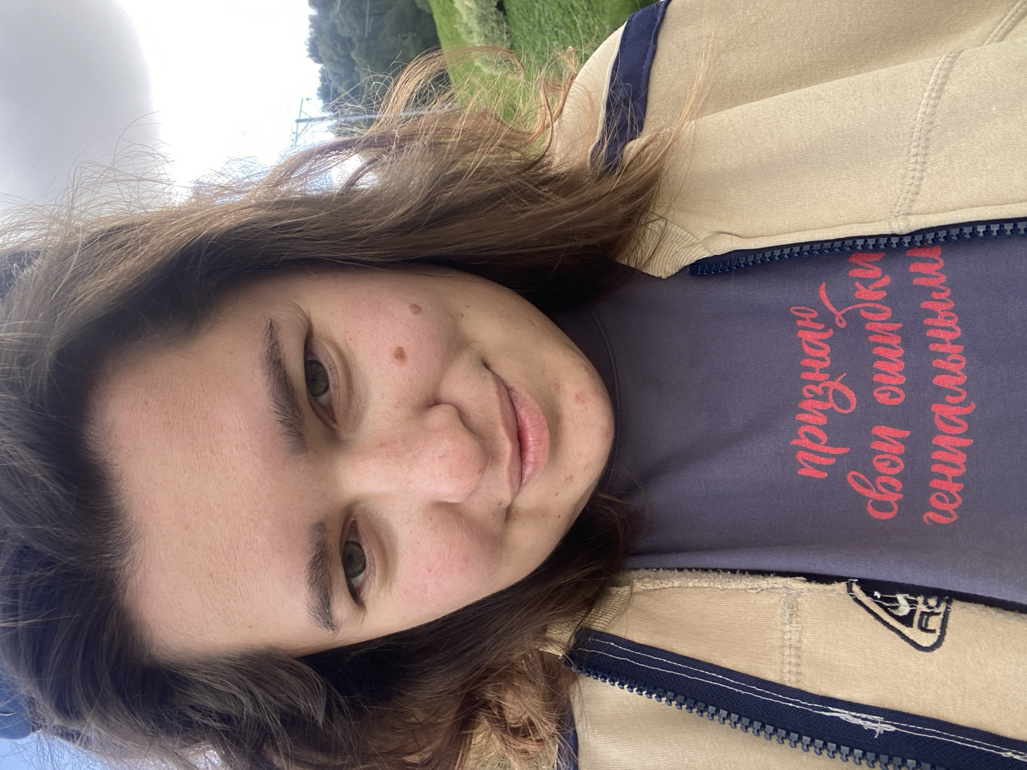 The image is a selfie of a young woman with medium-length brown hair, looking directly at the camera. She is wearing a tan jacket with blue trim and a dark purple t-shirt with pink Cyrillic lettering that reads "признаю свои ошибки гениальными" (I admit my mistakes are brilliant). She has a faint smile and visible pores and freckles on her face. The background is blurred but appears to be an outdoor setting with green foliage and a cloudy sky. The image does not contain any identifiable landmarks or features that would suggest a specific location within the city of Perm, Russia.
