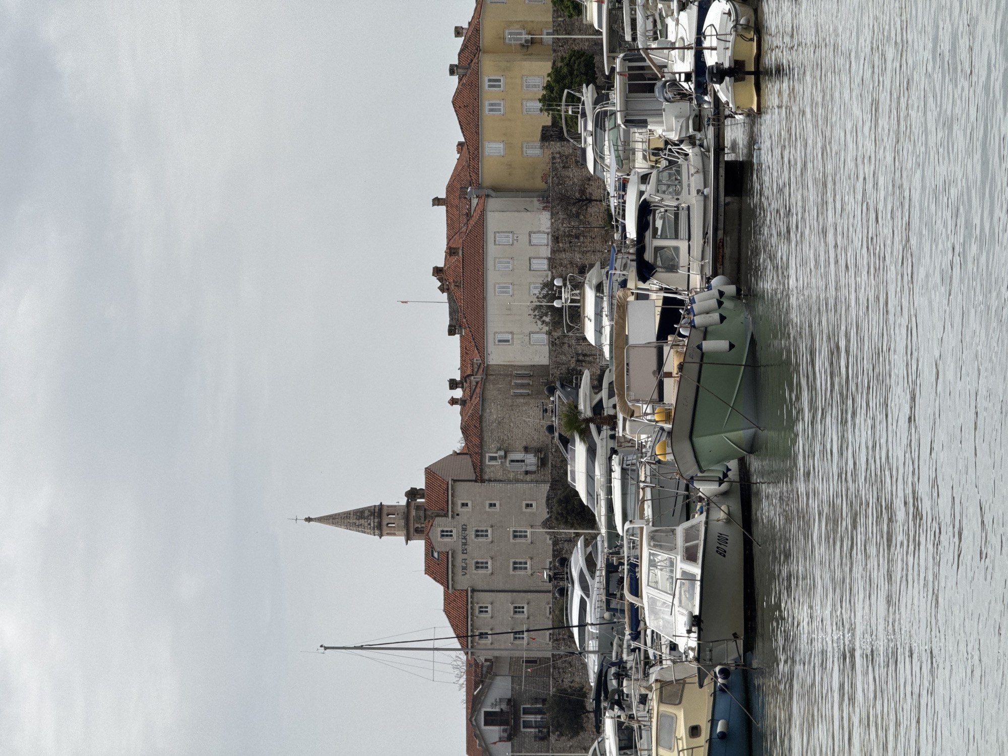 The image shows a marina in Budva, Montenegro, with various boats docked along the waterfront. A portion of the old city of Budva is visible in the background, featuring buildings with red-tiled roofs and a church spire. The sky is overcast. The water in the marina is calm, with reflections of the boats and buildings. The boats range in size and style, from small fishing boats to larger yachts. A sign on one building reads "VILA BALKAN." No people are visible.
