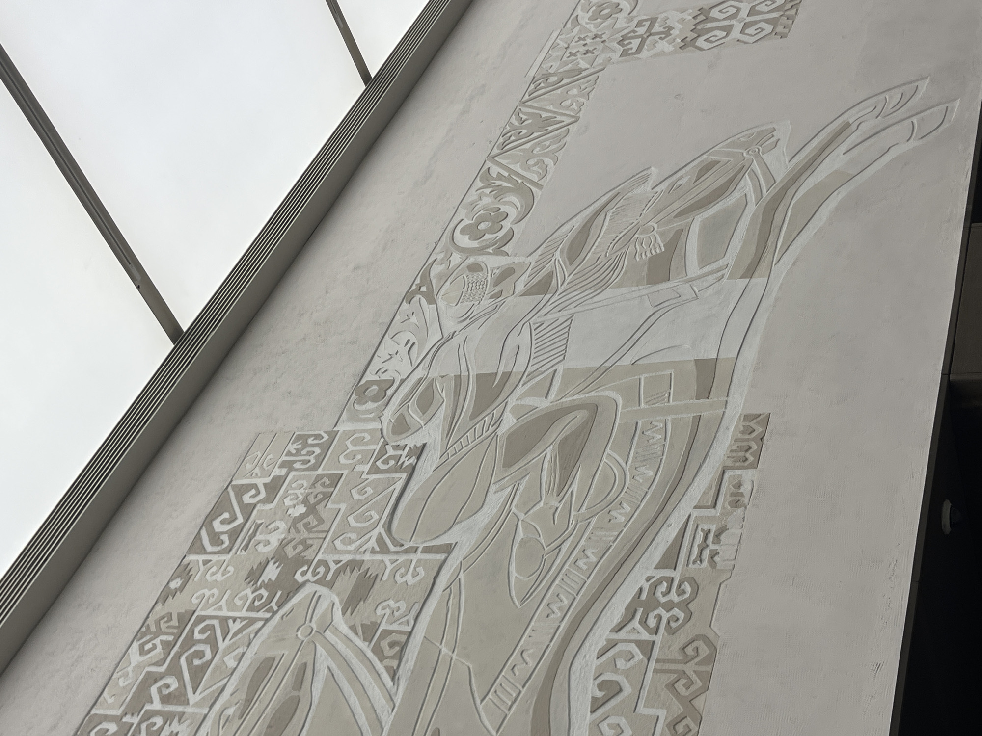 The image shows a close-up of a relief sculpture on a building's exterior in Almaty, Kazakhstan. The sculpture depicts a stylized scene with figures and geometric patterns in a monochromatic, low-relief style. The figures appear to be human or mythical, with flowing lines and intricate detailing. They are positioned on what looks like a stylized vehicle or a large animal, moving from left to right. The background is a plain, textured wall. Above the sculpture, there is a modern architectural element with horizontal slats and a bright, translucent panel, suggesting a roof or overhang. The overall impression is one of historical or cultural significance integrated into contemporary architecture.