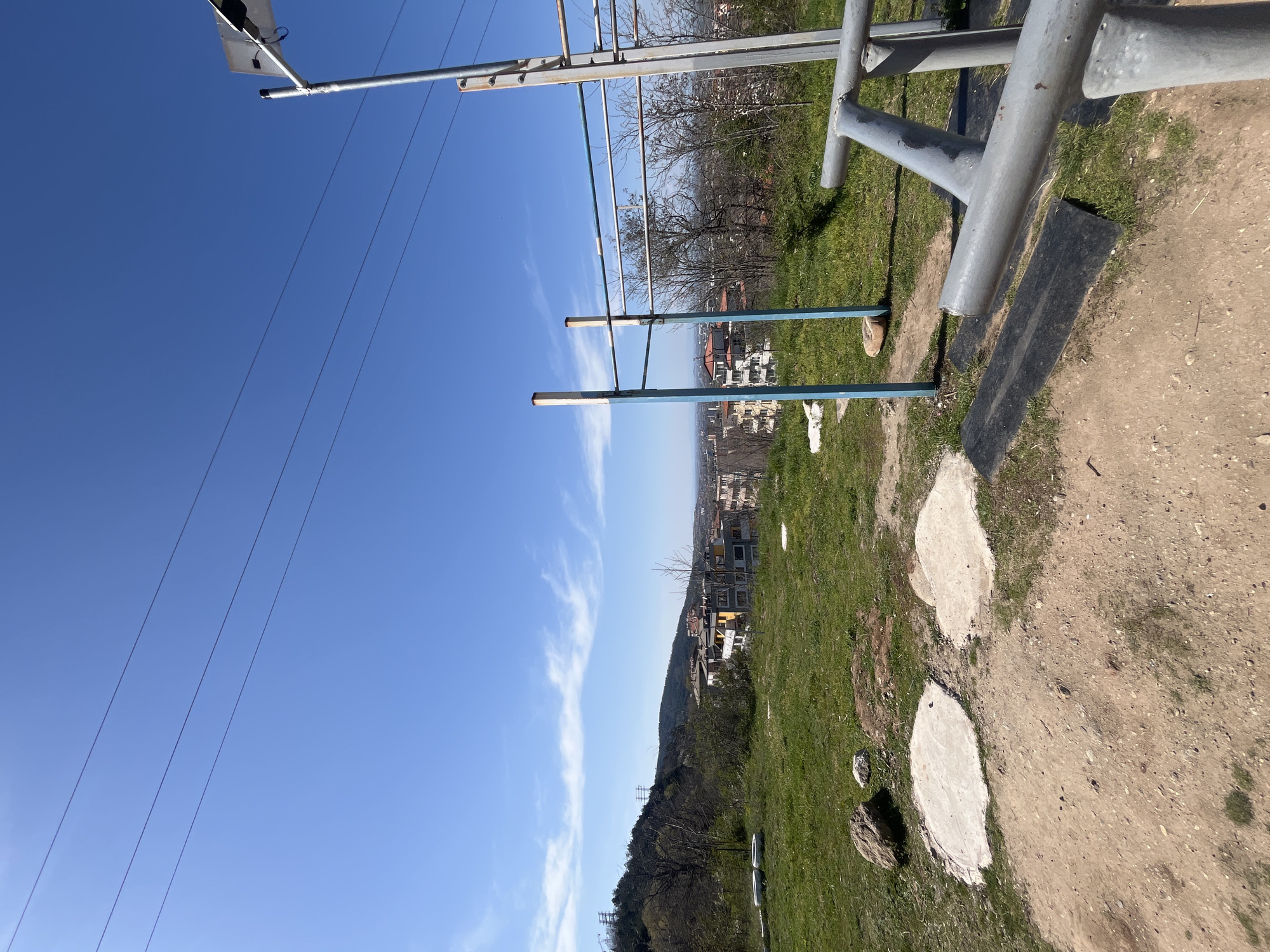 The image shows a park in Asenovgrad, Bulgaria on a sunny day with a clear blue sky. The scene is oriented uphill, with a partial view of the city in the background. In the foreground, there is an exercise area with parallel bars made of gray metal and blue rails. The ground is a mix of dirt, grass, and concrete stepping stones. Overhead power lines are visible against the sky. Trees line the hill beyond the park.