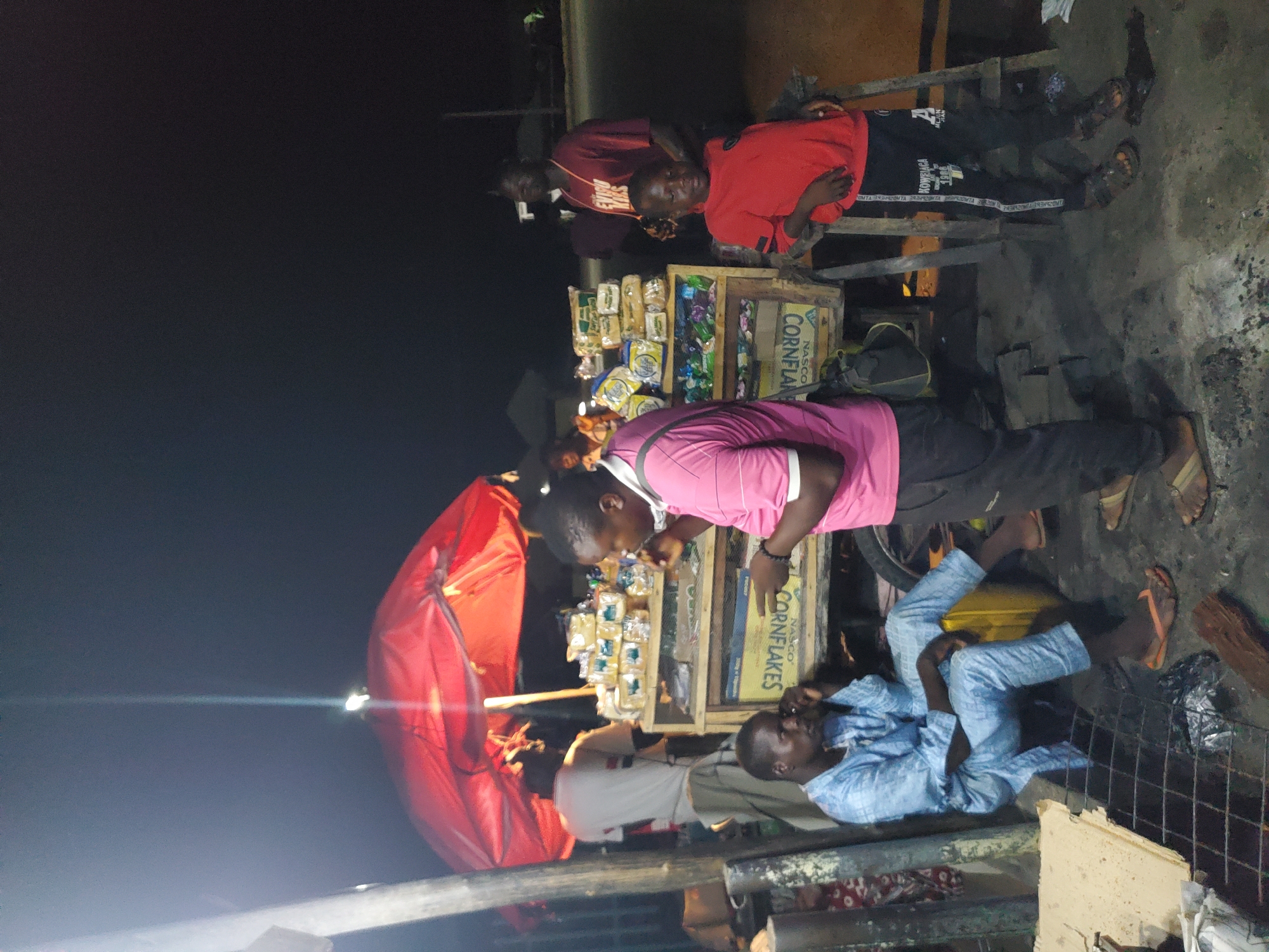 In the city of Jalingo, Nigeria, at night, a street vendor operates under a large red umbrella. The vendor's stall is wooden, displaying various packaged goods, including what appears to be cornflakes. A man in a light blue traditional outfit sits on a yellow container to the left of the stall, with his hand to his face. To the right of the stall, a man in a pink shirt and black pants stands, appearing to be in conversation with someone out of frame. A young boy in a red shirt and black pants stands to the right of the man in pink. In the background, another man in a maroon t-shirt is visible. The scene is illuminated by artificial light, casting shadows and highlighting the goods for sale. The ground is paved with dirt and debris.
