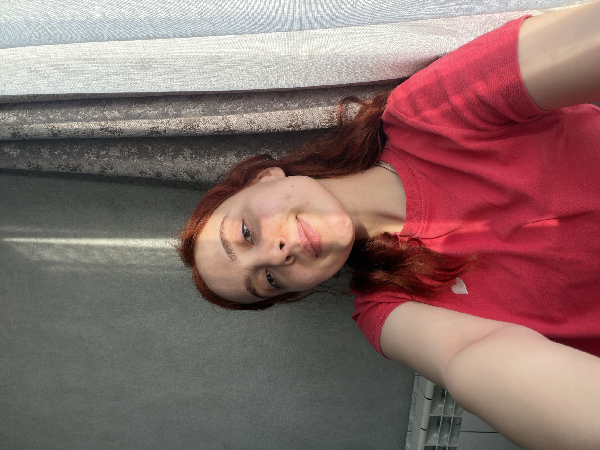 A selfie-style photograph depicts a fair-skinned person with red hair and blue eyes, wearing a red t-shirt. The person is oriented at an angle, with their head toward the upper left of the frame. Behind the person is a grey wall and a layered, textured curtain. The location cannot be determined from the image.
