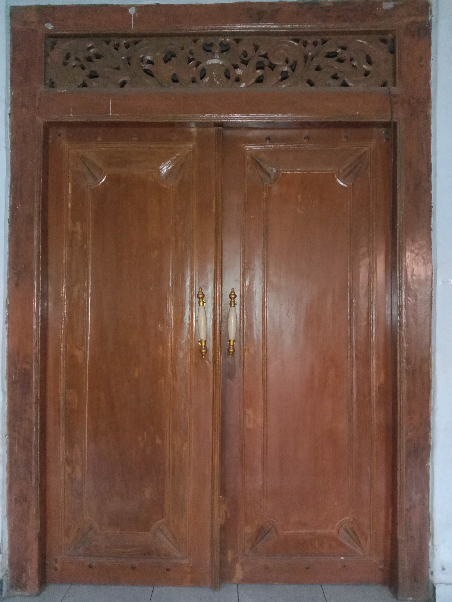 The image shows a pair of wooden double doors, likely an entrance to a building. The doors are made of polished brown wood with decorative carving at the top of the frame, featuring floral or organic motifs. Each door has two vertical panels with a beveled edge and an inward-facing, pointed decorative element near the bottom. Two brass and cream-colored handles are visible, one on each door, positioned centrally. The doors are set within a wooden frame. The surrounding environment is dimly lit, with hints of a white wall to the right and a tiled floor or threshold at the bottom. The image appears to have been taken indoors or in a covered outdoor space. The scene is static, with no people or activity depicted. The location is specified as Pohsangit Leres, Indonesia.