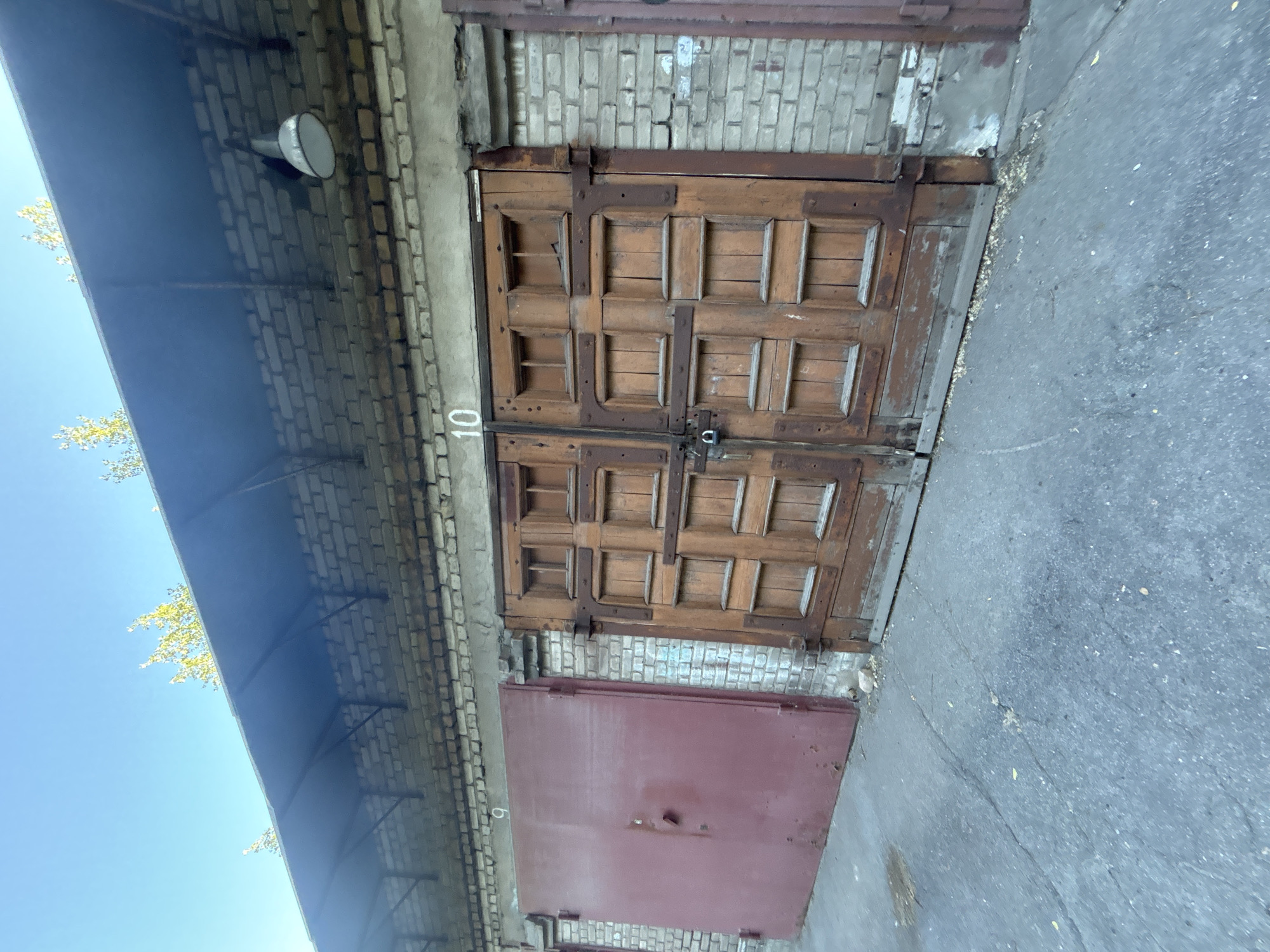 The image shows an exterior view of a building with two doors. The primary subject is a large, weathered wooden double door with a rustic design, featuring multiple square panels and dark metal hardware. To the left and below this wooden door is a smaller, plain, reddish-brown metal door. The building is constructed of light-colored brick. Above and to the left of the doors, a blue awning extends outwards, partially obscuring some of the brickwork and a white outdoor light fixture. The scene appears to be outdoors, with a clear blue sky visible to the left. A grey asphalt surface is in the foreground, suggesting a driveway or pathway. The overall impression is one of an older, functional building, possibly a garage or storage area, with signs of wear and tear on the doors. The lighting suggests it is daytime, with clear weather. The number "10" is visible in white stencil font on the brick wall to the left of the wooden door.