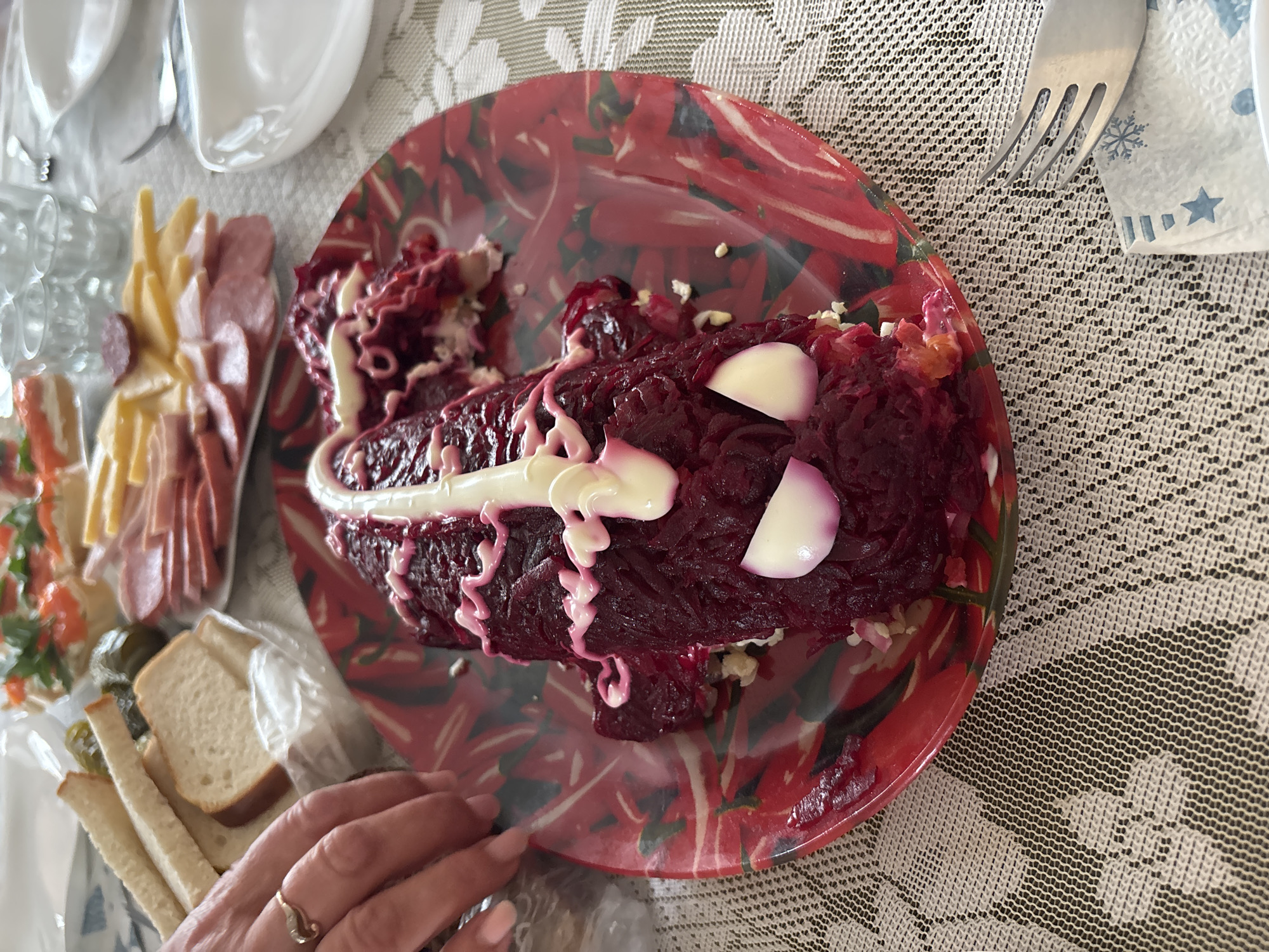 The image shows a dining table in what appears to be a home setting. The table is covered with a white lace tablecloth. On the table is a red patterned plate with a serving of a dark red salad, possibly "Herring Under a Fur Coat", decorated with mayonnaise and sliced eggs. Next to the plate is a fork and a napkin with a snowflake and star design. To the left of the plate, there's a platter with various food items, including cheese, sliced meats, pickles, and slices of white bread. A woman's hand is near the bread. There are no indications of the location being Krasnoyarsk, Russia in the image. The environment appears to be indoors.